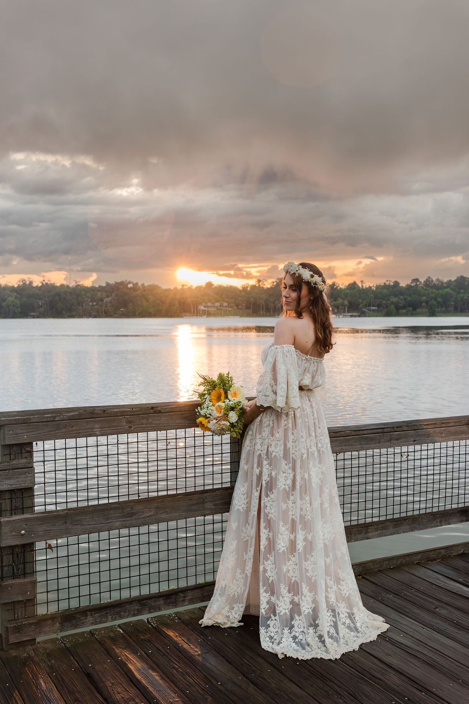 Bride in lace off-shoulder gown holds yellow bouquet on wooden pier at sunset over calm lake.