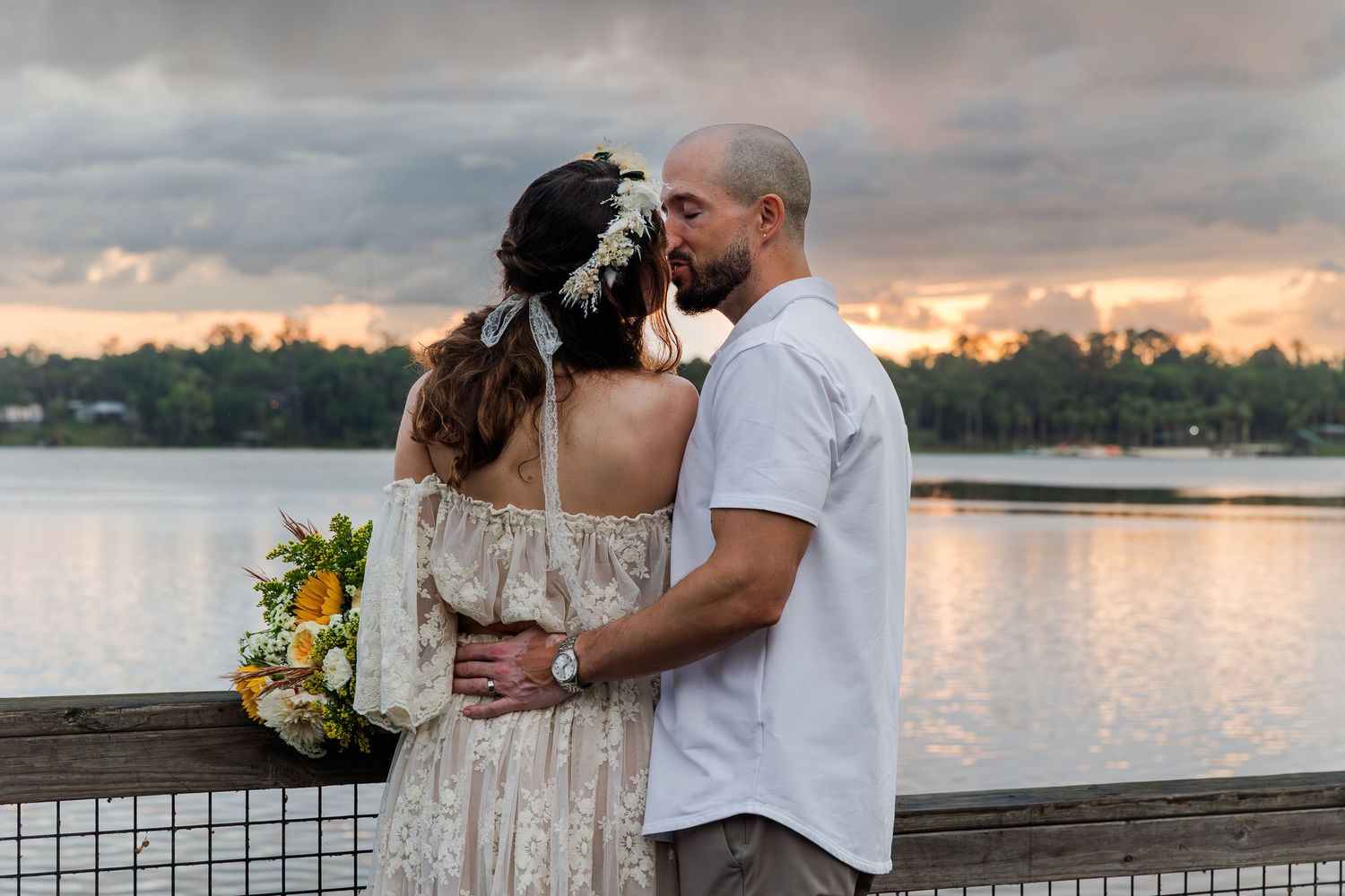 Couple embraces on a lakeside dock at sunset, bride holding a colorful bouquet under a cloudy sky.