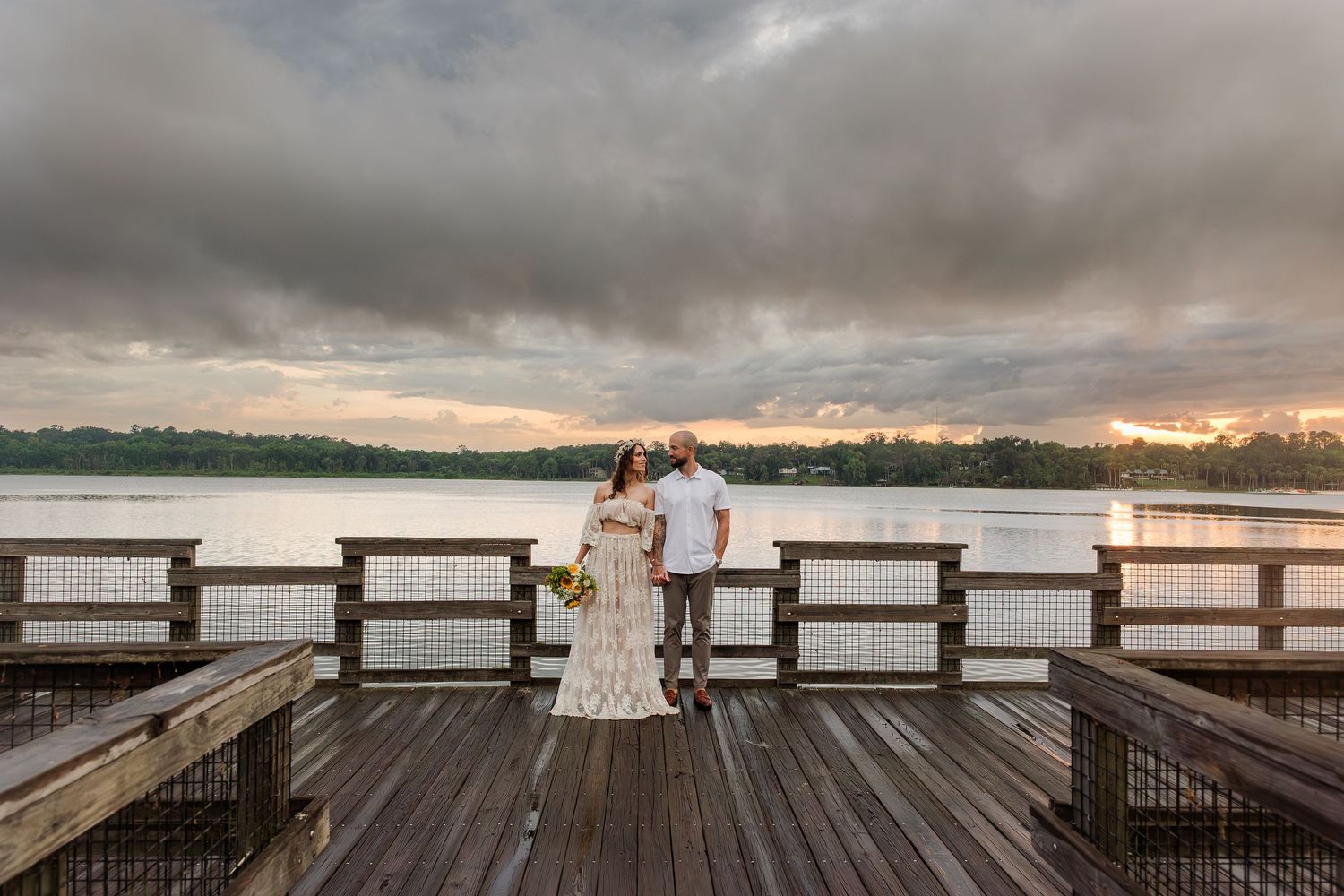 Couple in wedding attire poses on wooden pier overlooking calm lake at sunset with dramatic cloudy sky.