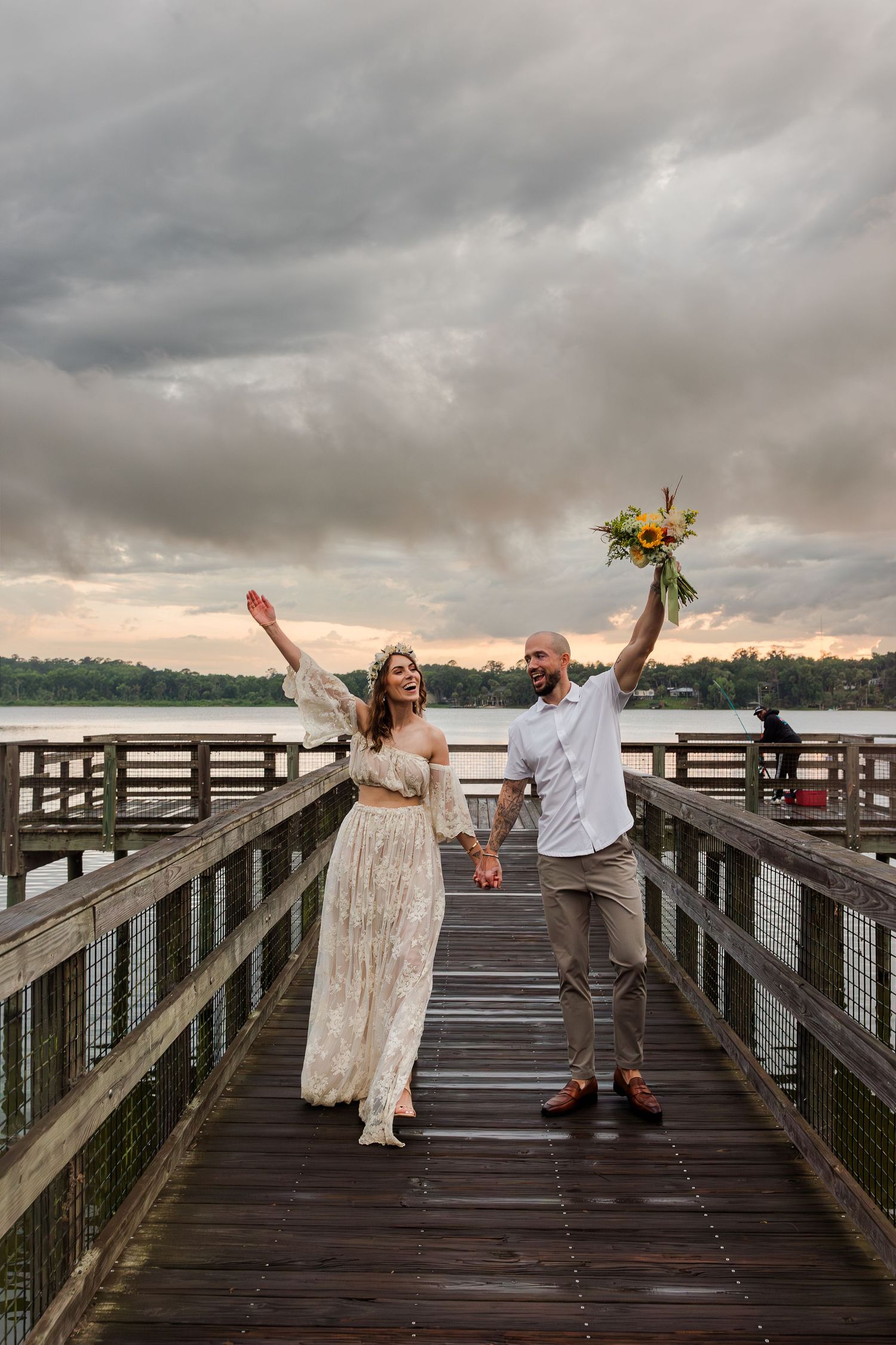 Joyful newlyweds celebrate on a wooden pier, bride in lace dress holding bouquet aloft under dramatic cloudy sky.