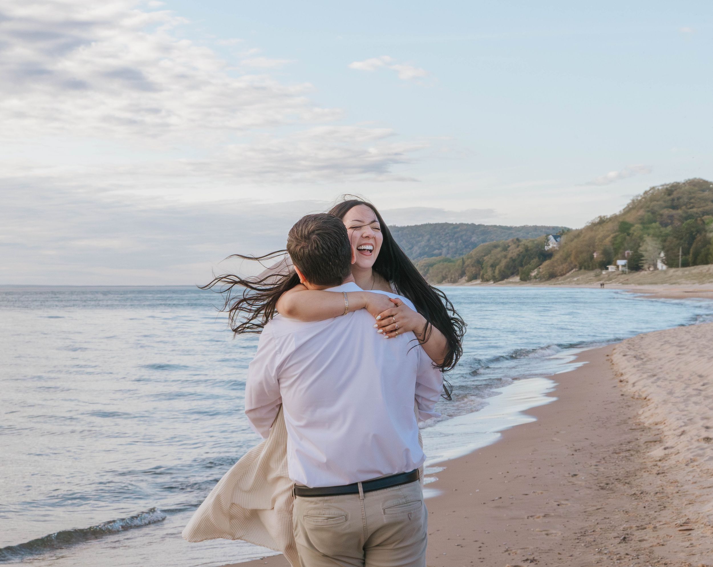 Happy couple embracing and spinning on a sandy beach shore with ocean waves and cloudy sky backdrop.