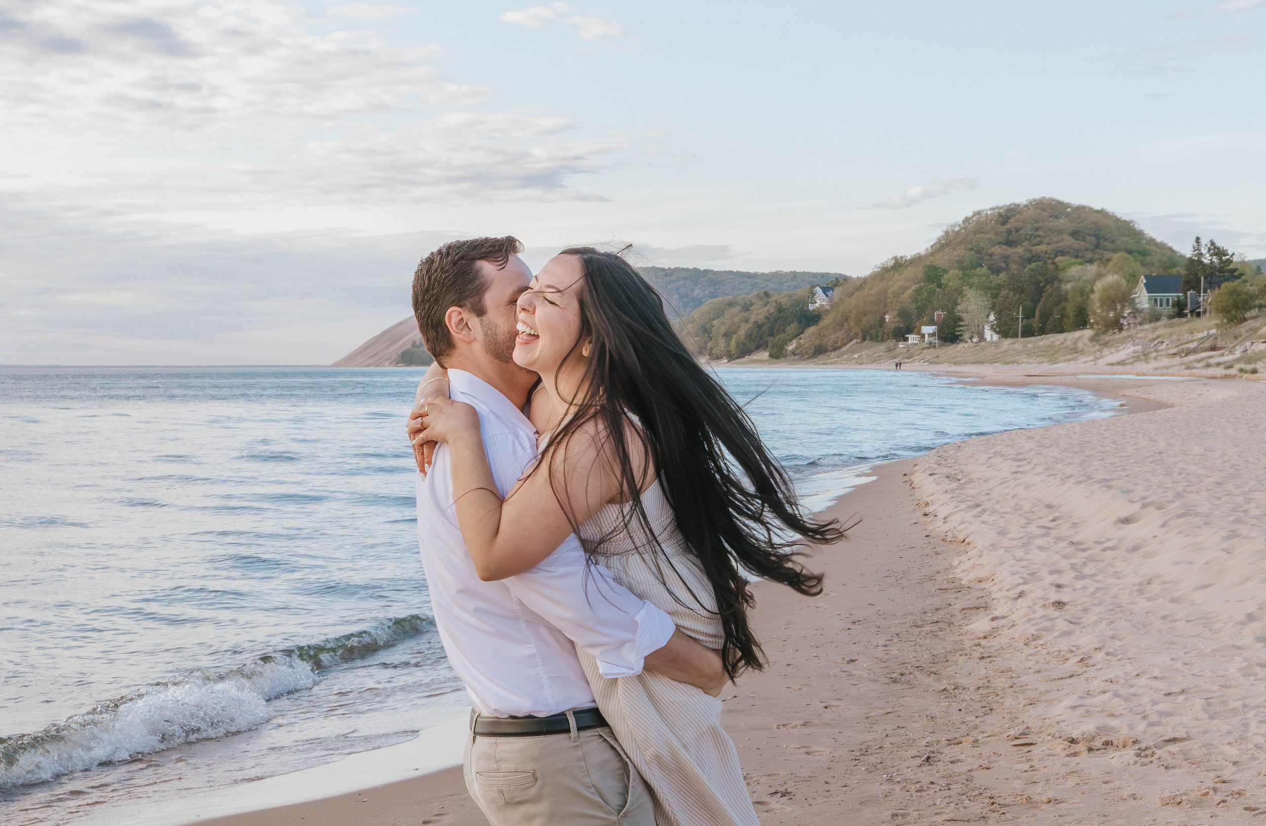 Couple embracing and laughing on a sandy beach with ocean waves and green hills in the background.