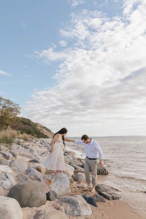 Couple walking hand-in-hand on a rocky shoreline with ocean waves and a cloudy sky backdrop.