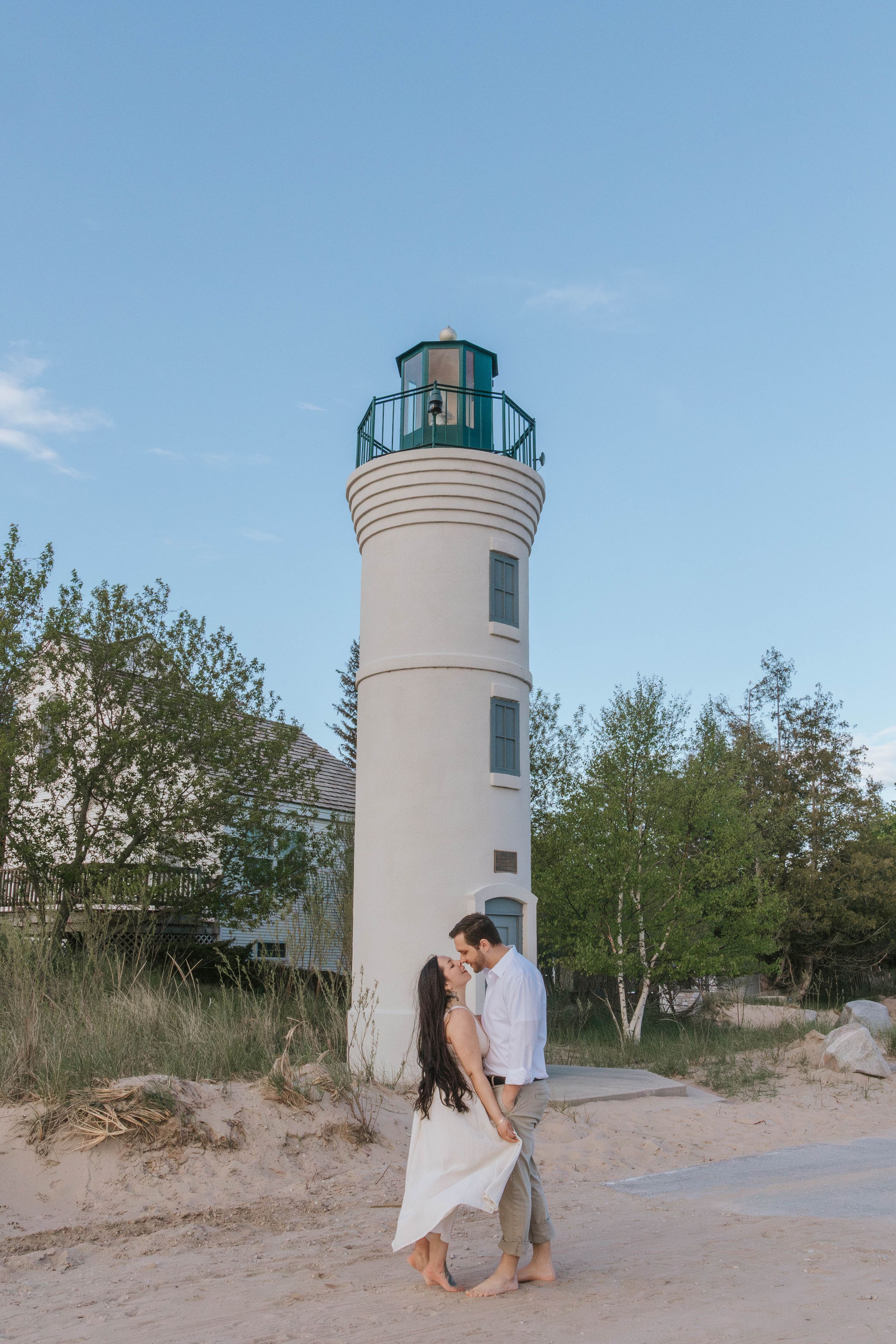 Couple sharing a romantic kiss on sandy beach in front of a white lighthouse surrounded by green trees.