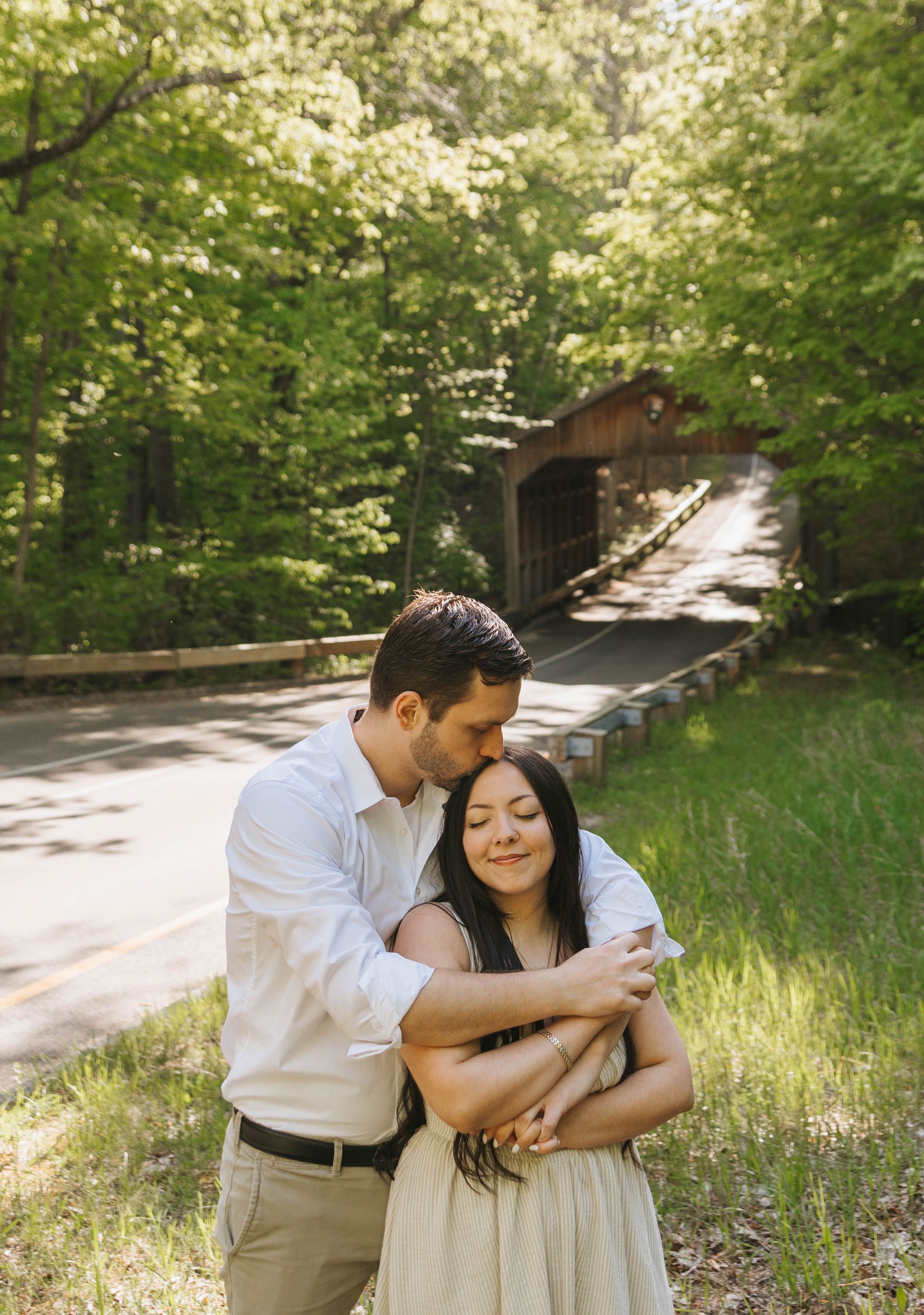 A couple embraces near a covered bridge surrounded by lush green trees during a romantic outdoor engagement session.