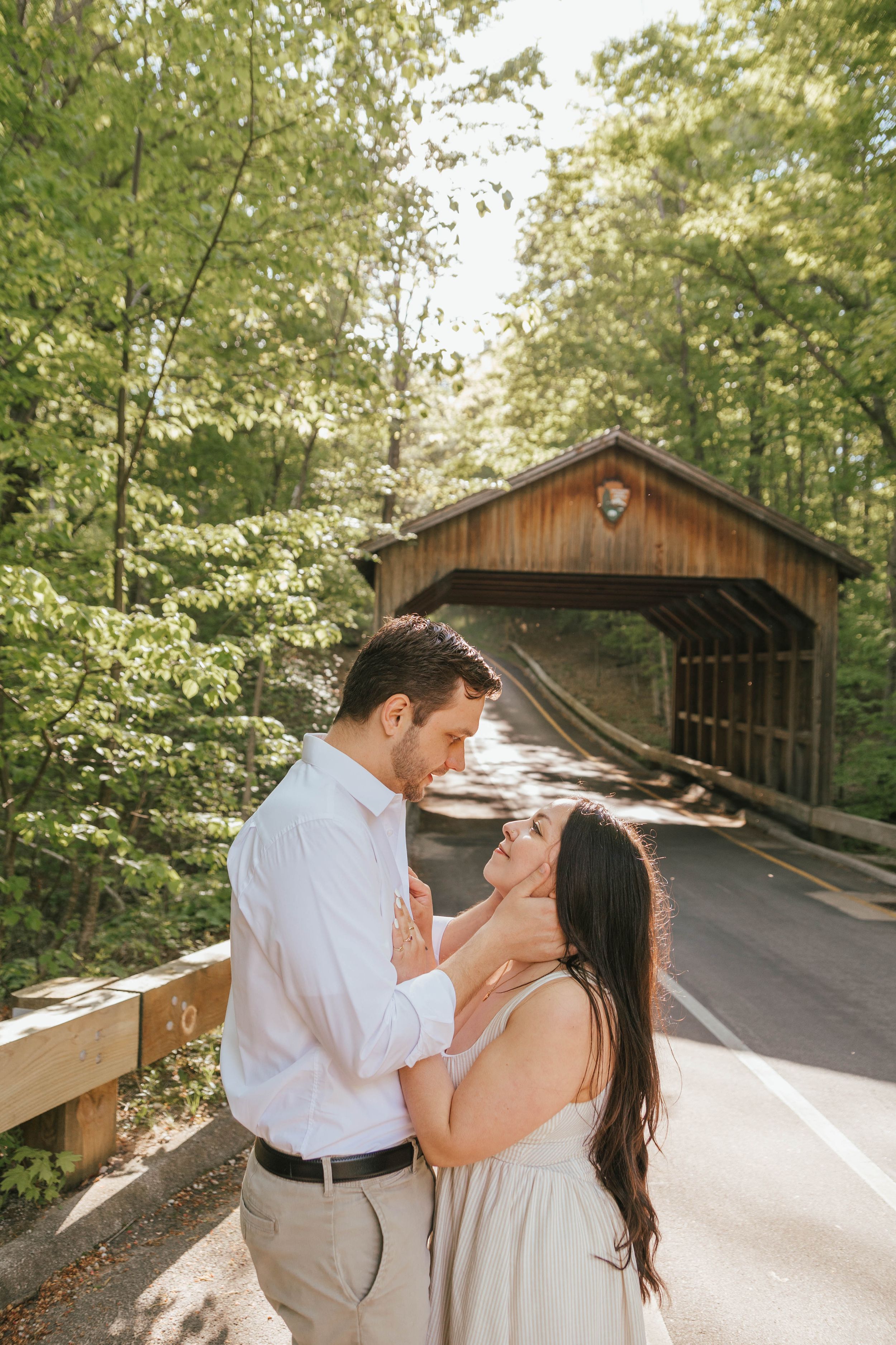 Couple embracing near a wooden covered bridge surrounded by lush green trees on a sunny day.