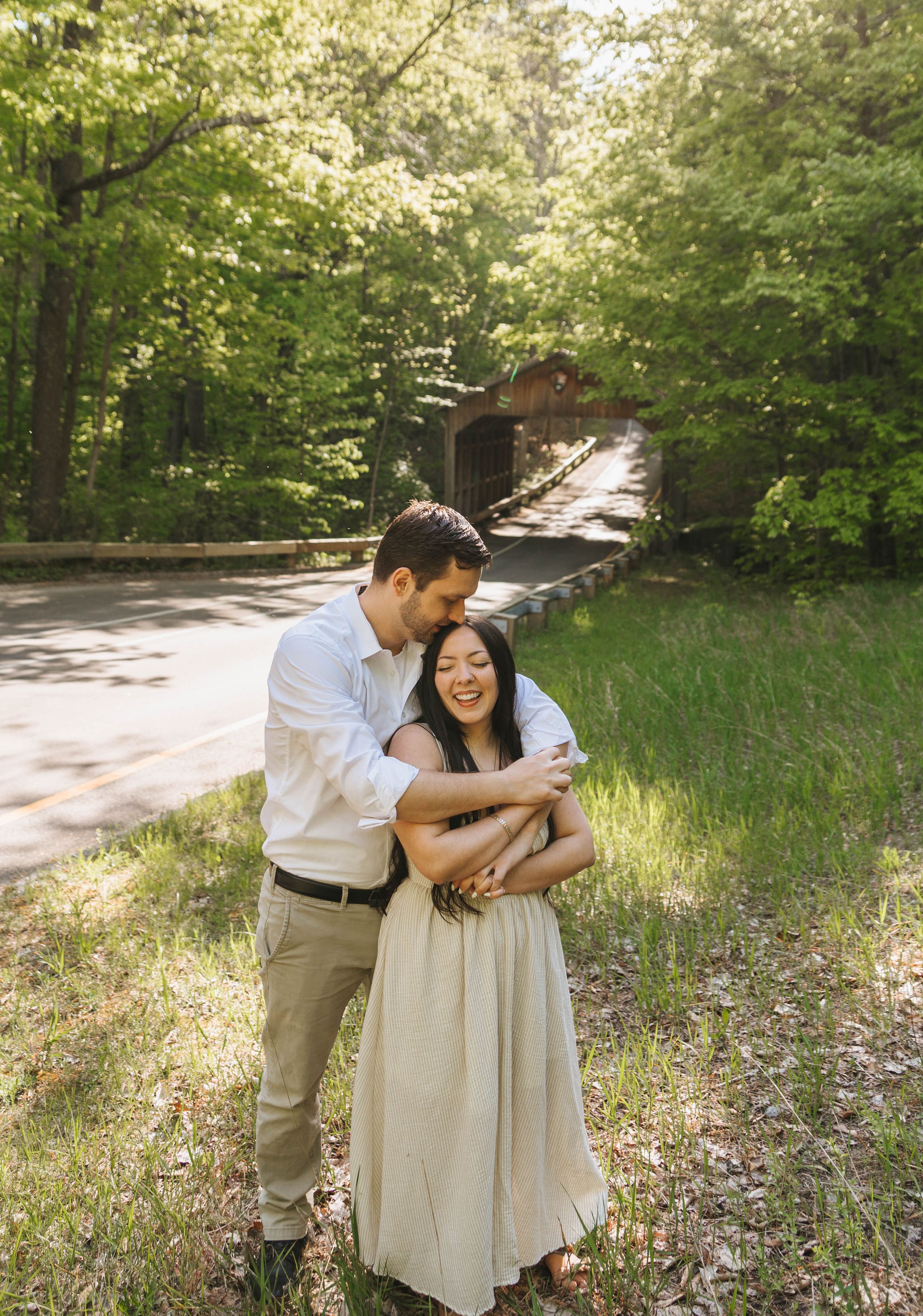 Couple embracing outdoors near a covered bridge surrounded by lush green trees during an engagement photo session.