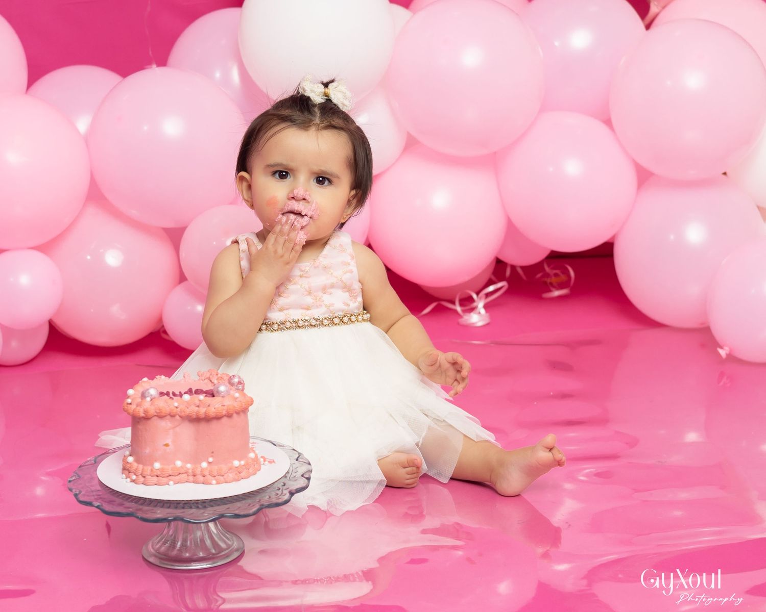 Baby girl in pink dress smashes first birthday cake surrounded by pink balloons in a professional photo shoot.
