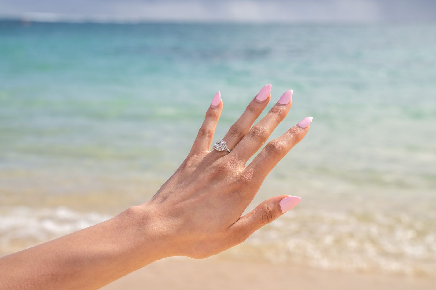A hand with pink French tip nails wearing a diamond engagement ring posed against a tropical beach background.