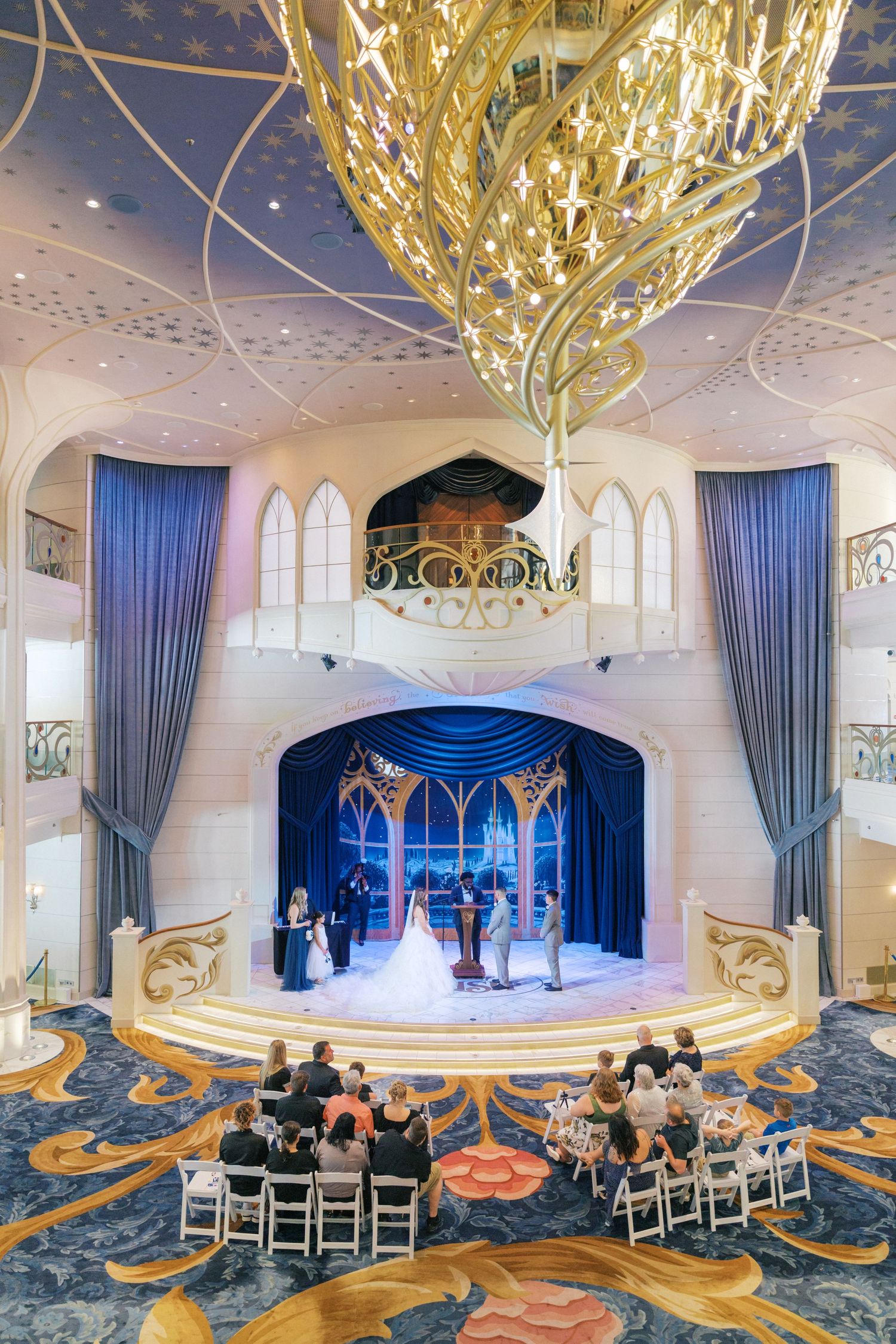 Elegant Disney cruise ship wedding ceremony in grand atrium with blue drapes, gold chandelier, and seated guests.