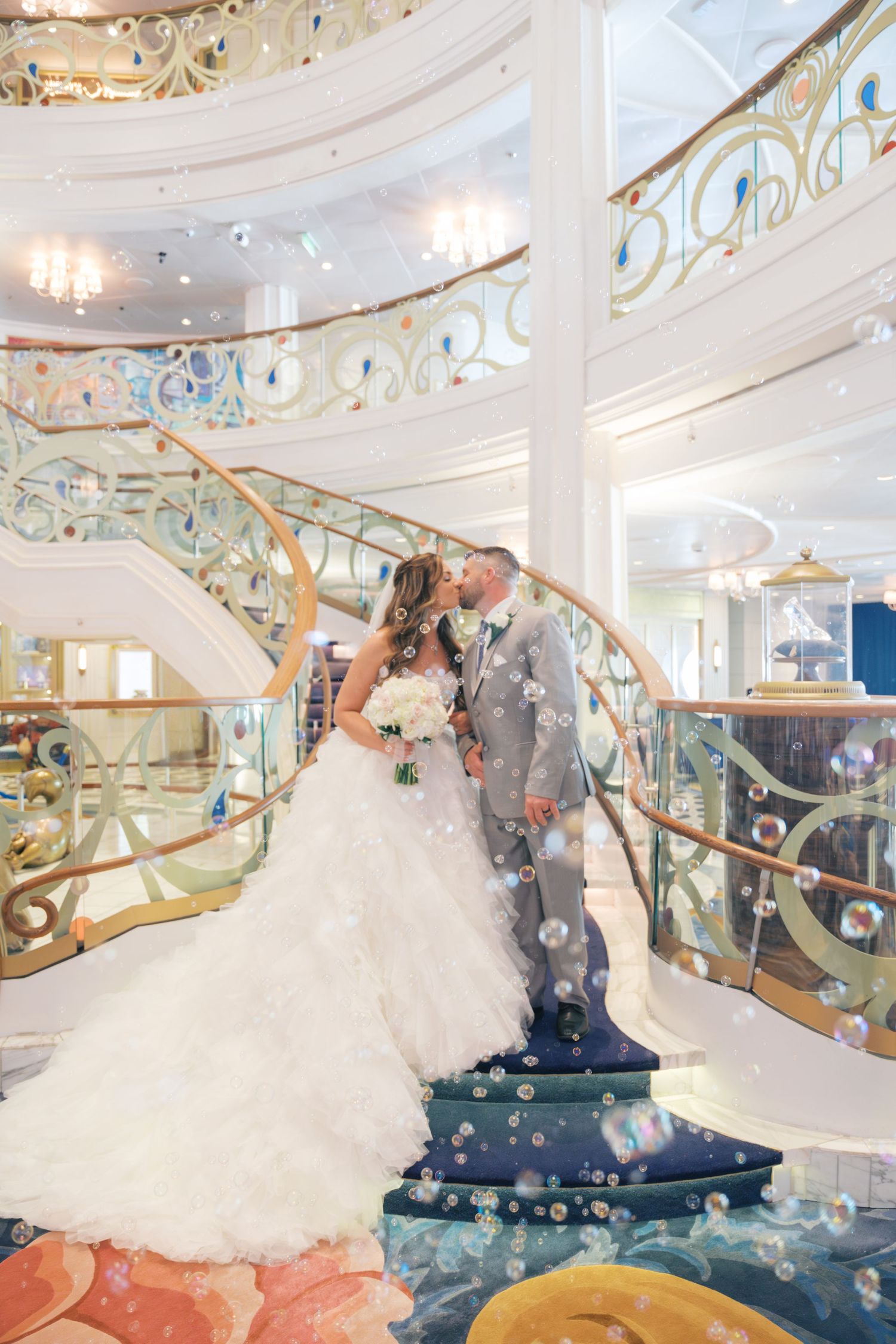 Bride and groom share a kiss on an elegant cruise ship staircase surrounded by floating bubbles.