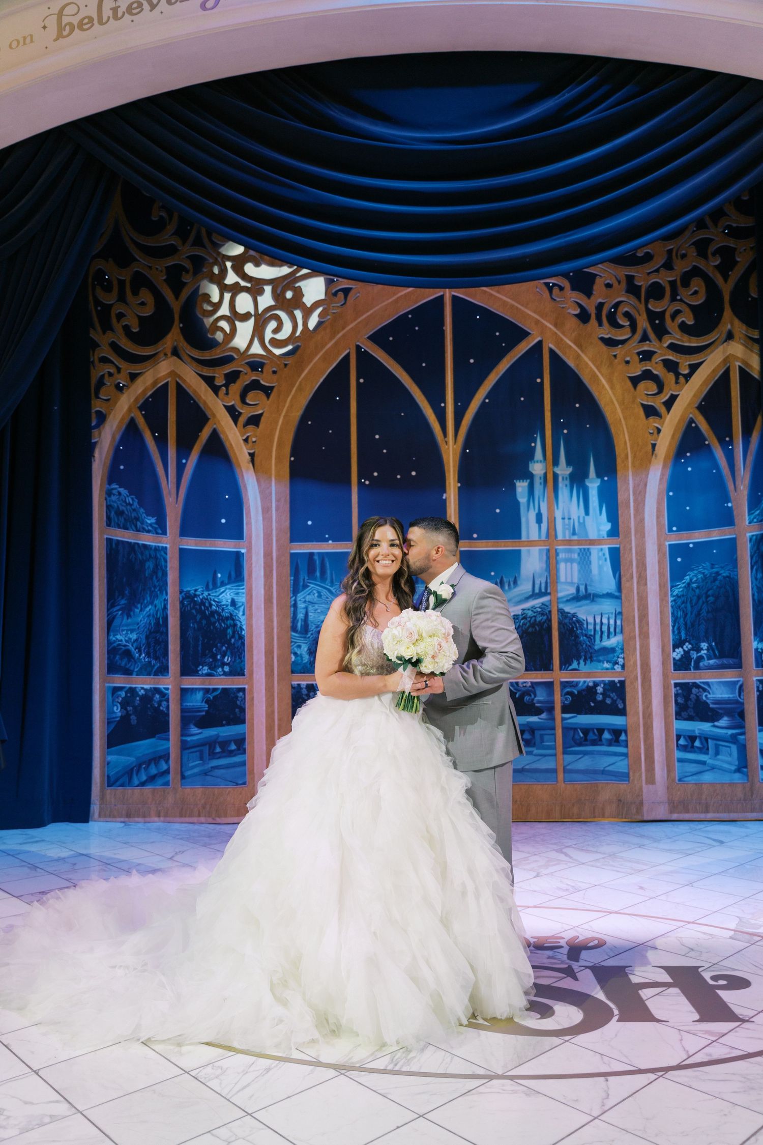 Bride and groom share romantic kiss on Disney cruise ship stage with enchanted castle backdrop.