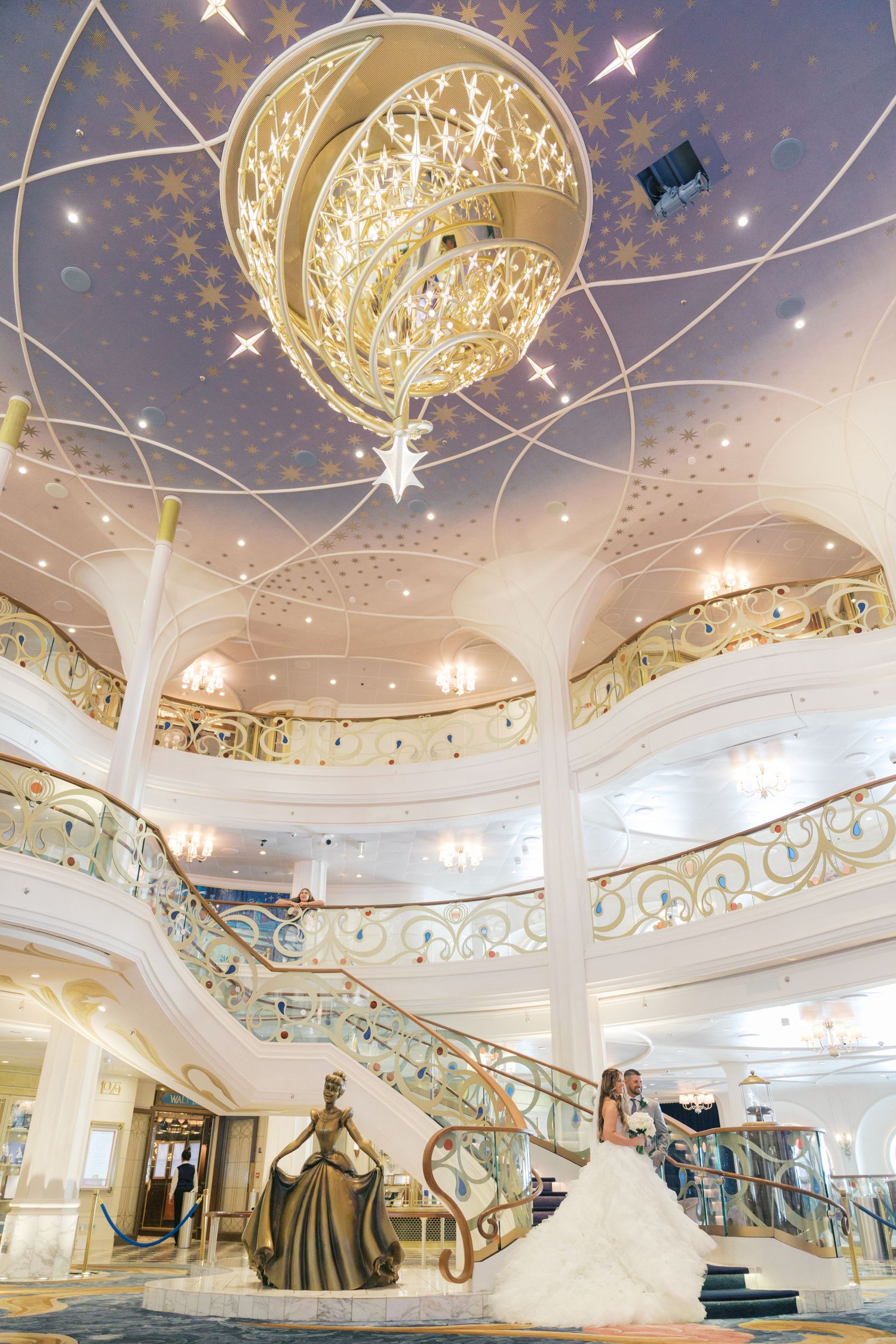 Bride poses near Cinderella statue in Disney cruise ship grand atrium with ornate staircase and chandelier.