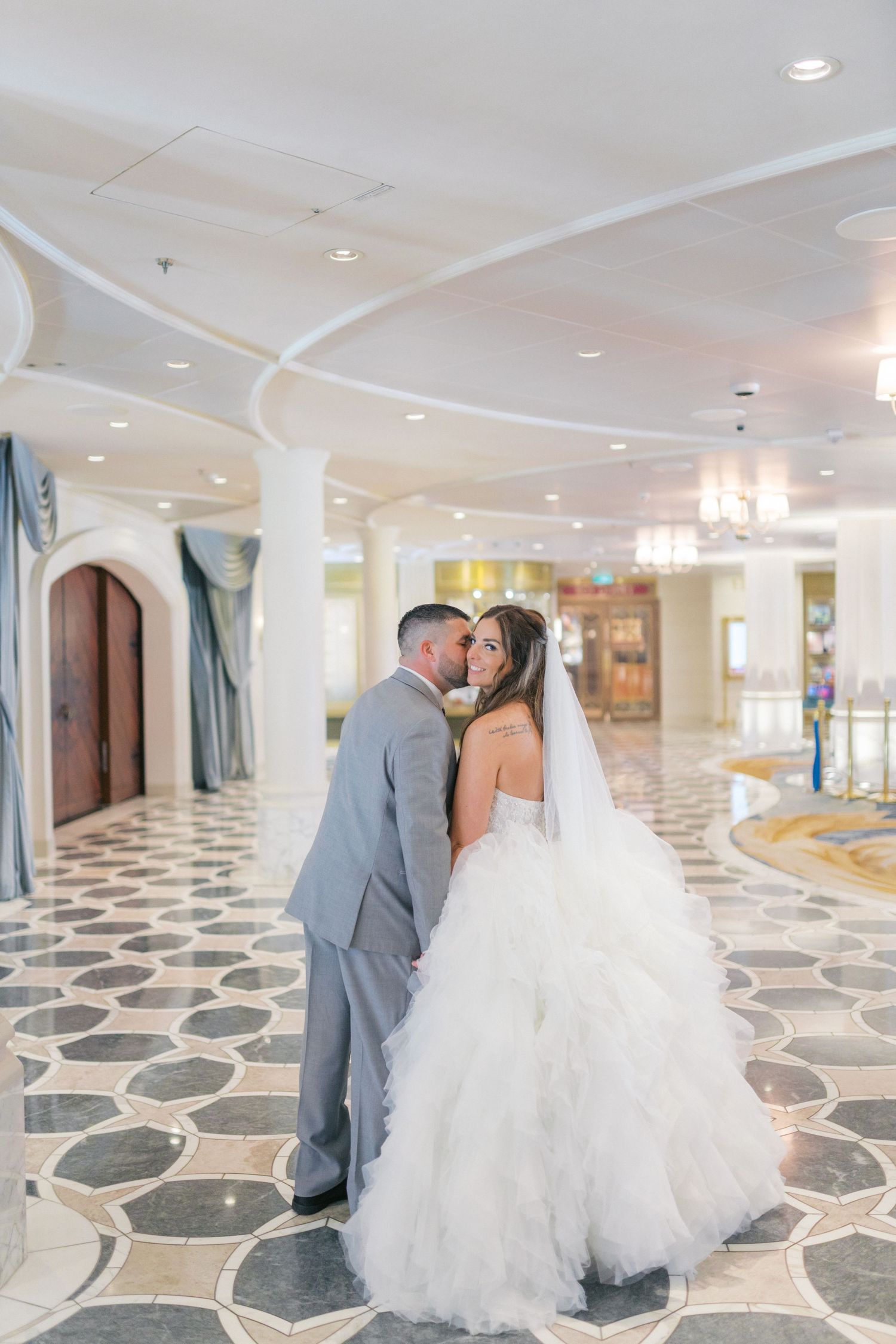 Bride and groom share a romantic kiss in an elegant marble-floored wedding venue lobby, bride wearing a ruffled white ballgown.