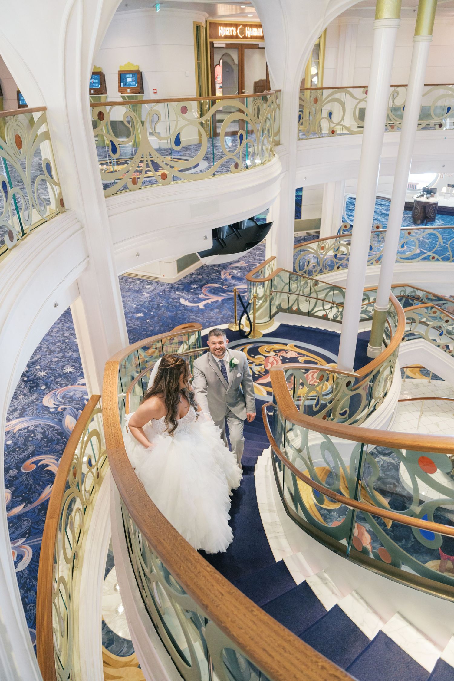 Bride and groom walk hand-in-hand down a grand spiral staircase on a luxury cruise ship.
