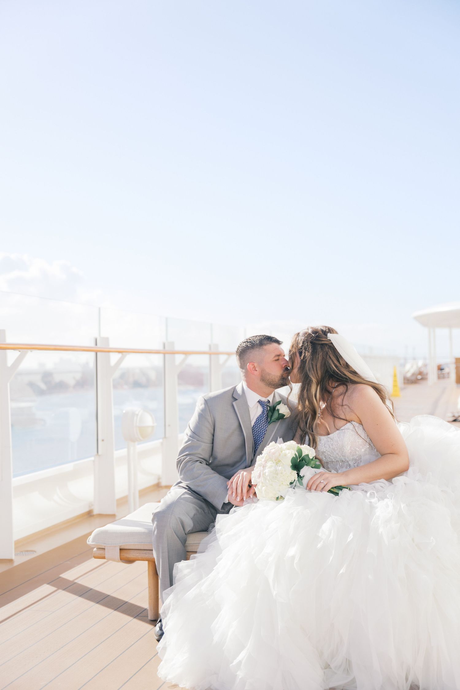 Bride and groom share a romantic kiss on a sunlit cruise ship deck with ocean views.