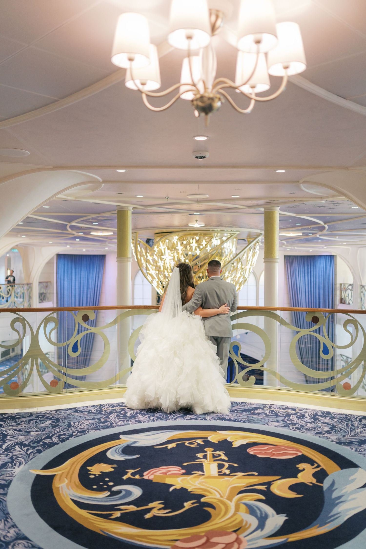 Bride and groom stand together on an ornate cruise ship staircase landing beneath an elegant chandelier.