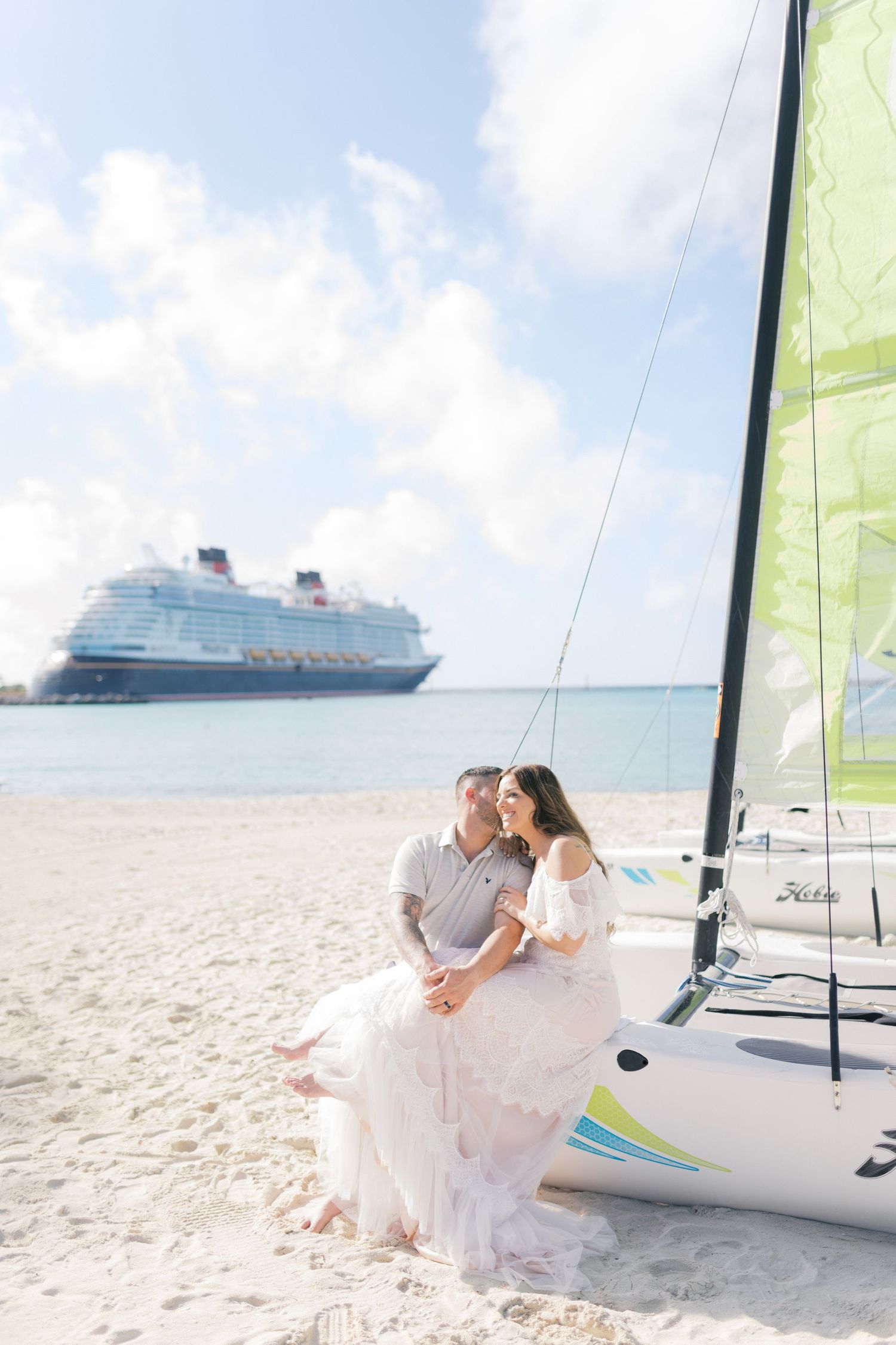 Couple sitting together on a sandy beach beside a Hobie Cat sailboat with a Disney cruise ship in the background.
