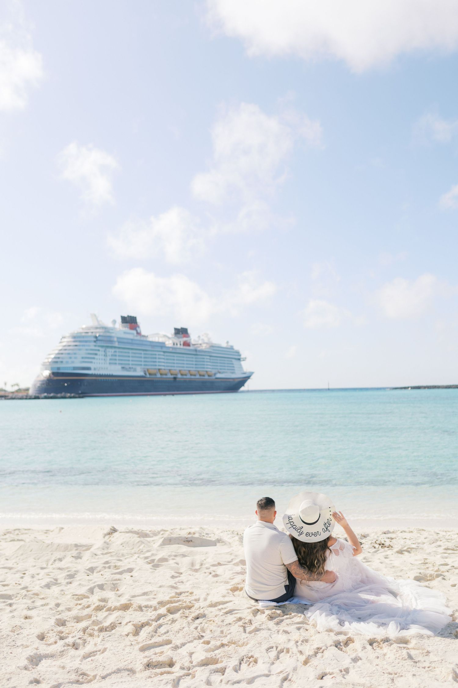 Couple in wedding attire sitting on white sand beach gazing at a Disney cruise ship on turquoise waters.