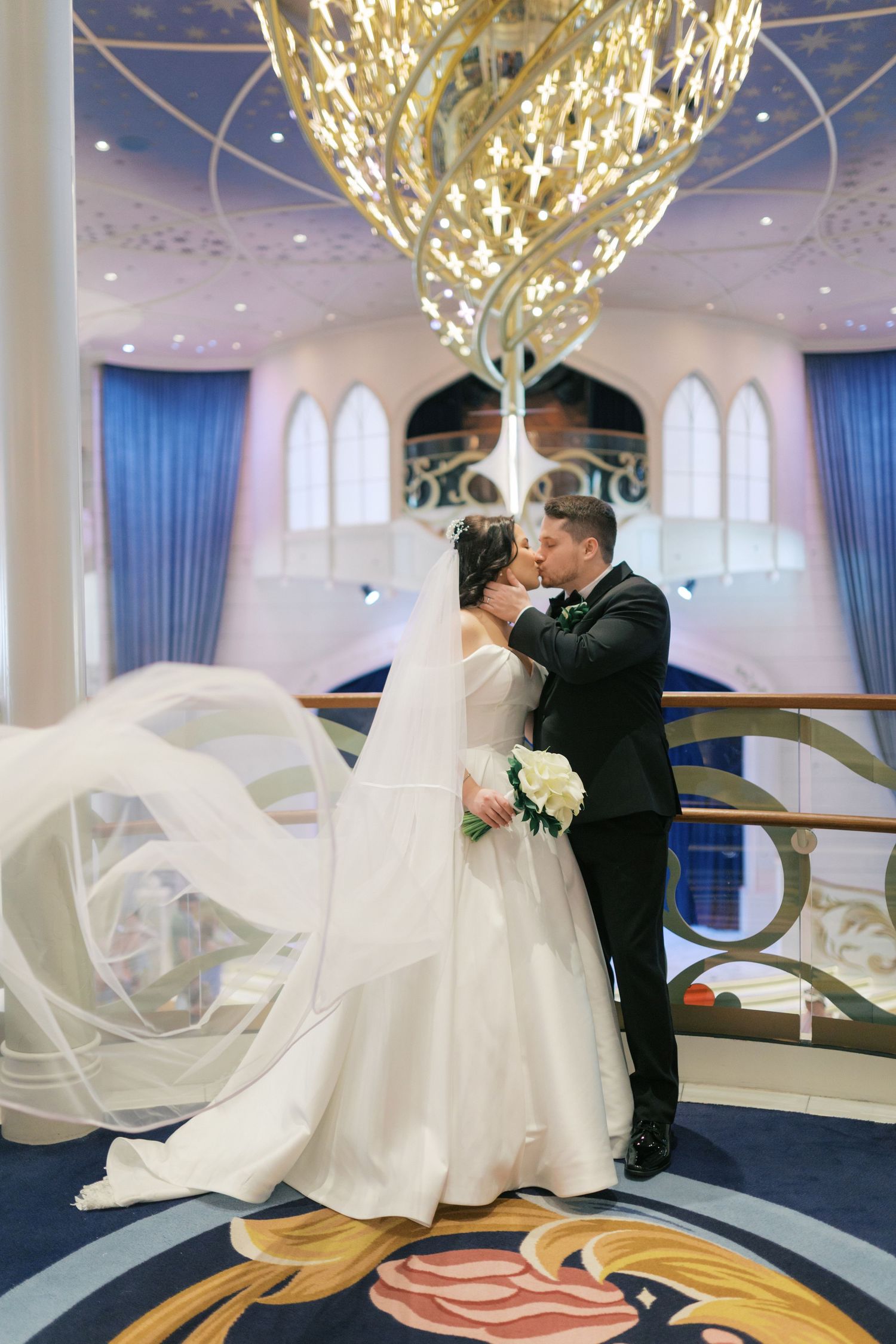 Bride and groom share a kiss under a grand chandelier in an elegant ballroom, with the wedding veil flowing dramatically.