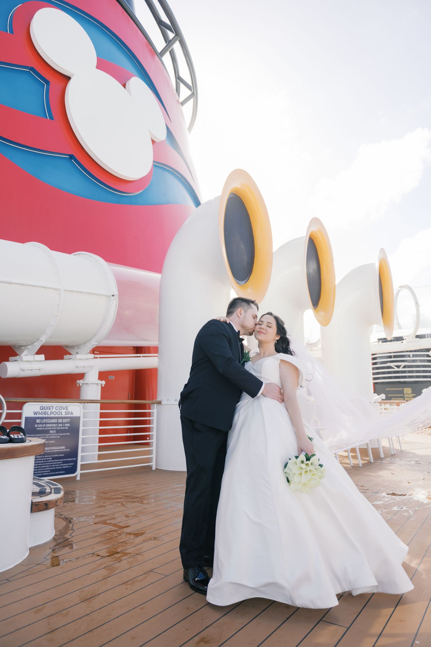 Bride and groom share a romantic kiss on the deck of a Disney cruise ship, with Mickey Mouse funnels in the background.