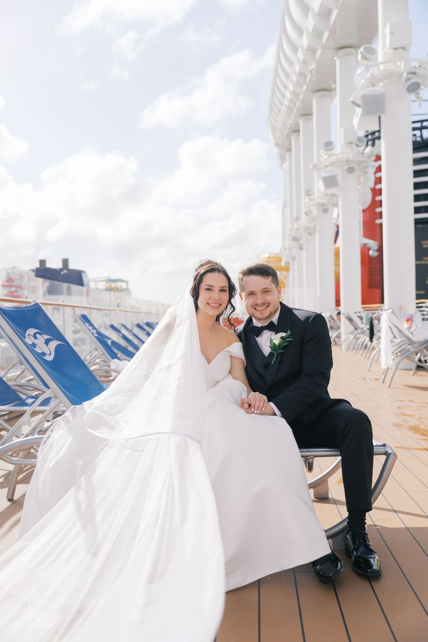 Bride and groom sitting together on cruise ship deck chairs, wearing wedding attire with flowing white gown.