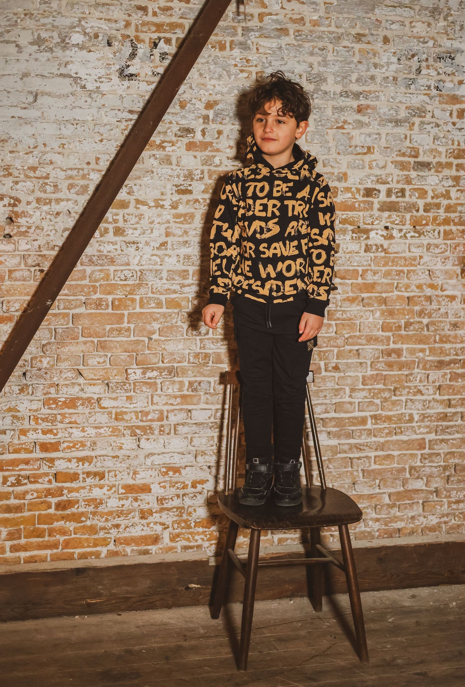 A young boy stands on a wooden chair wearing a black and gold text-print hoodie and black pants against a brick wall.