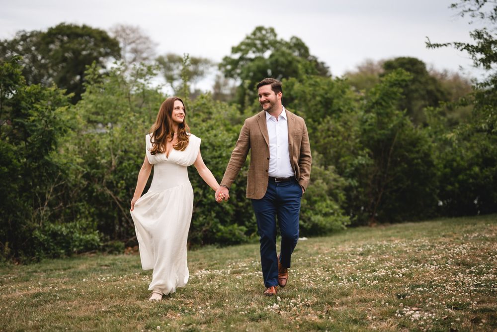 A couple holds hands walking through a green meadow, woman in white dress, man in tan blazer and navy pants.