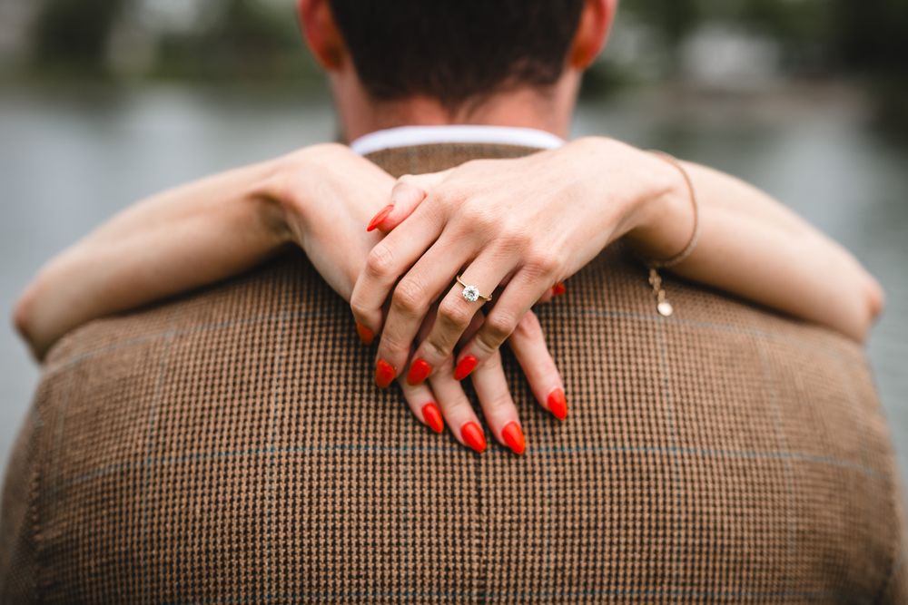 Woman with red nails and engagement ring embracing man in brown tweed jacket from behind.