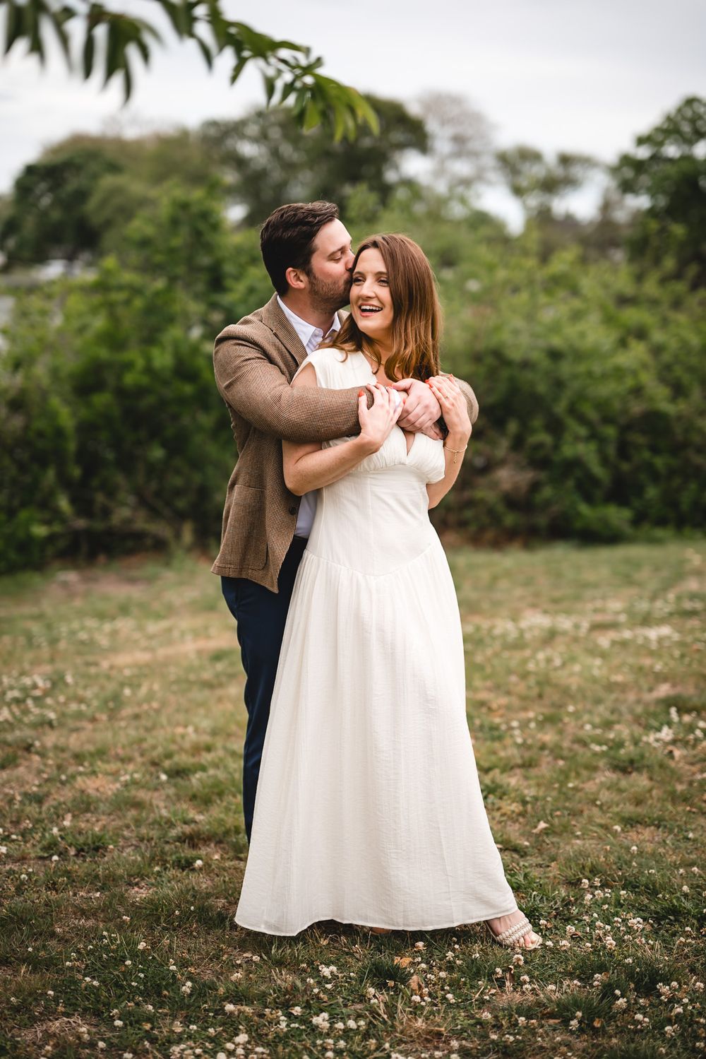 Couple embracing outdoors, man in brown jacket kissing woman in white wedding dress cheek, green trees background.