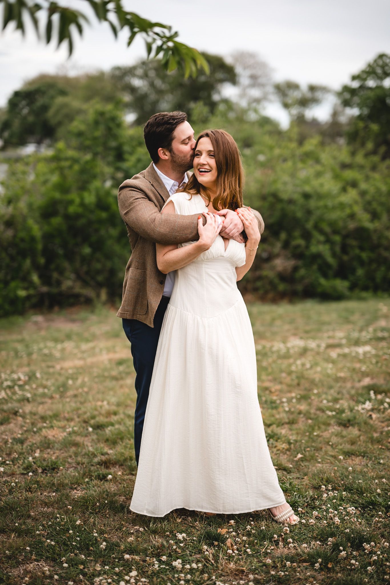 Couple embracing outdoors, man in brown jacket kissing woman in white wedding dress cheek, green trees background.