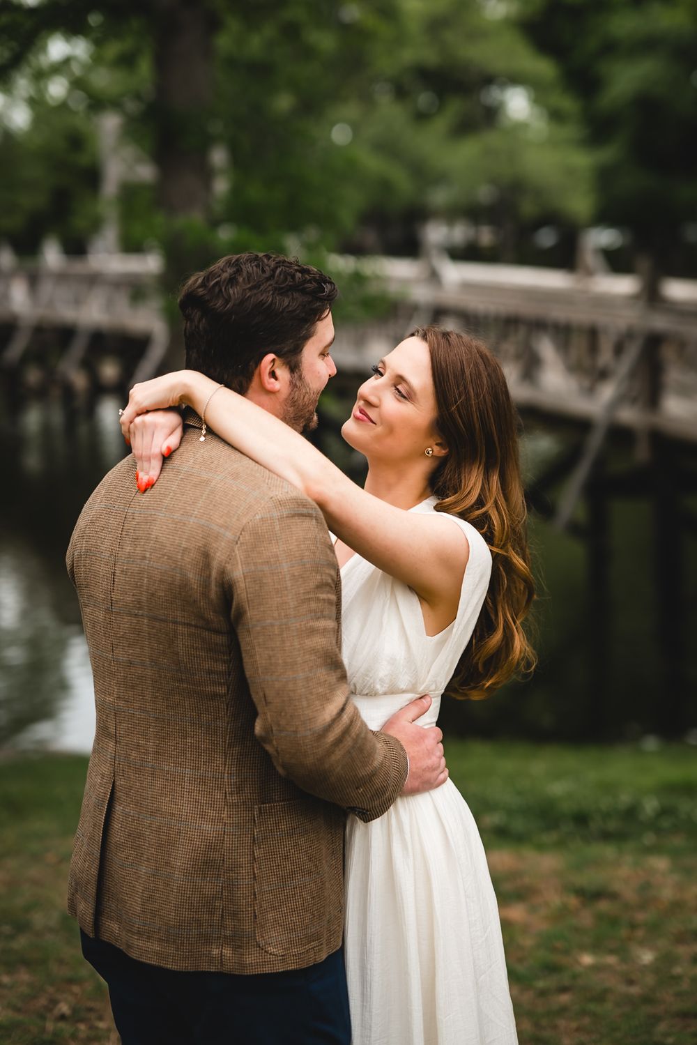 Couple embracing outdoors near a wooden bridge, woman in white dress smiling at man in brown tweed jacket.