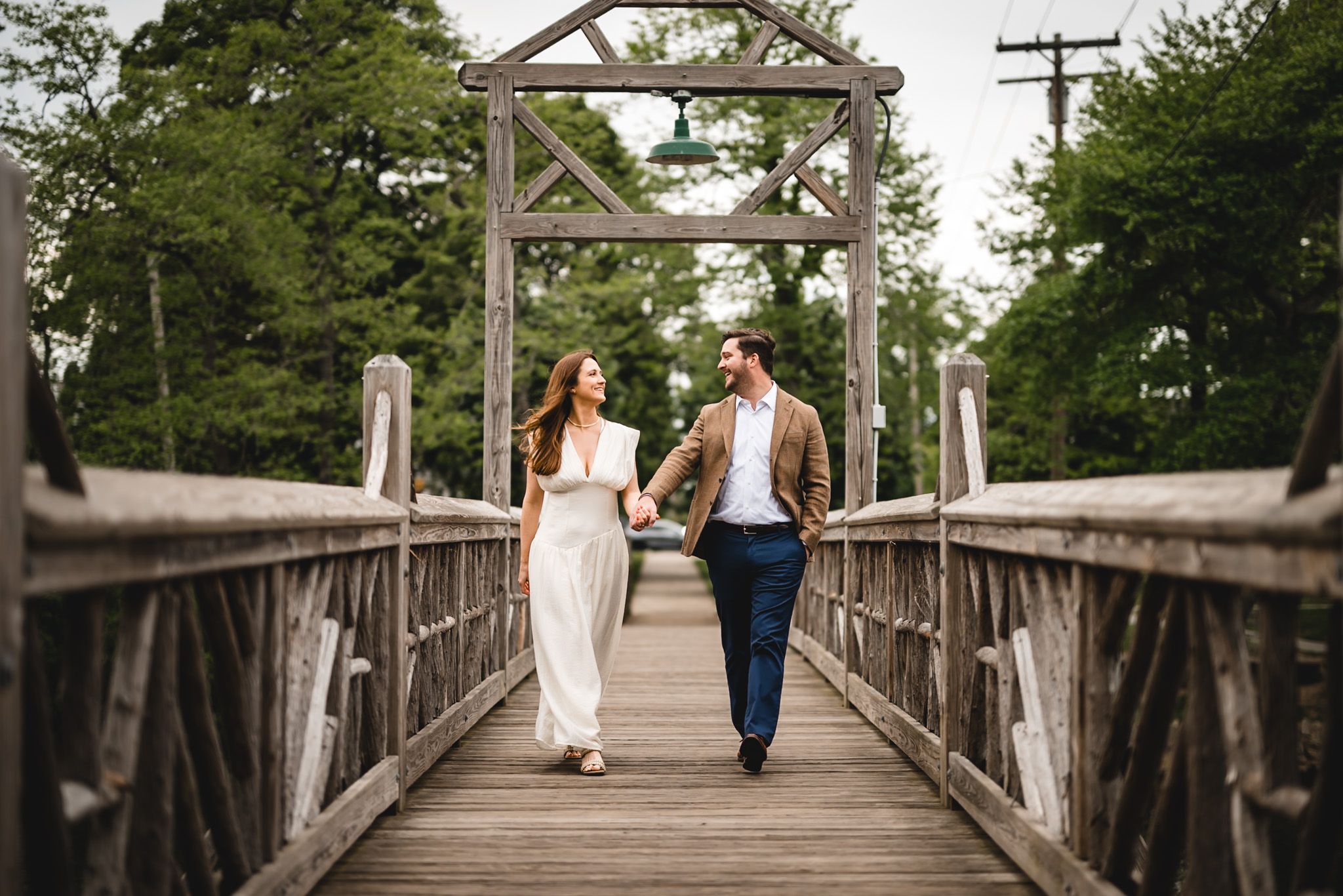 Couple holding hands walking on a wooden bridge surrounded by lush green trees during an outdoor engagement session.
