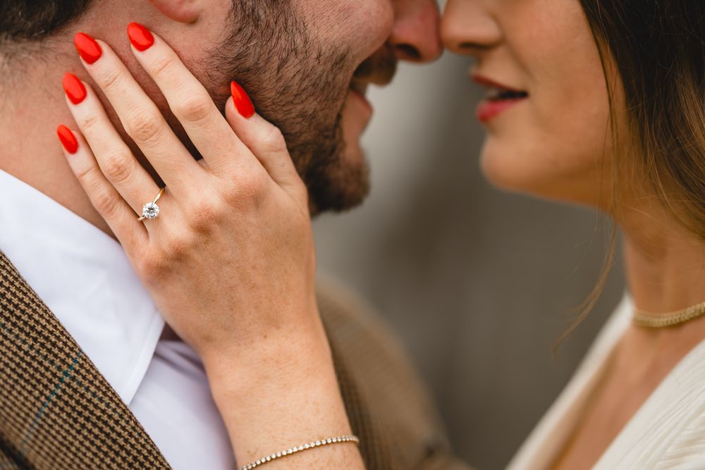 Close-up of couple almost kissing, woman showing diamond engagement ring with red nails and gold bracelet.
