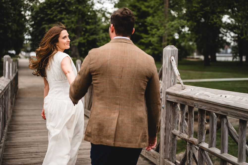 Bride and groom walking hand-in-hand on a wooden bridge surrounded by lush green trees.
