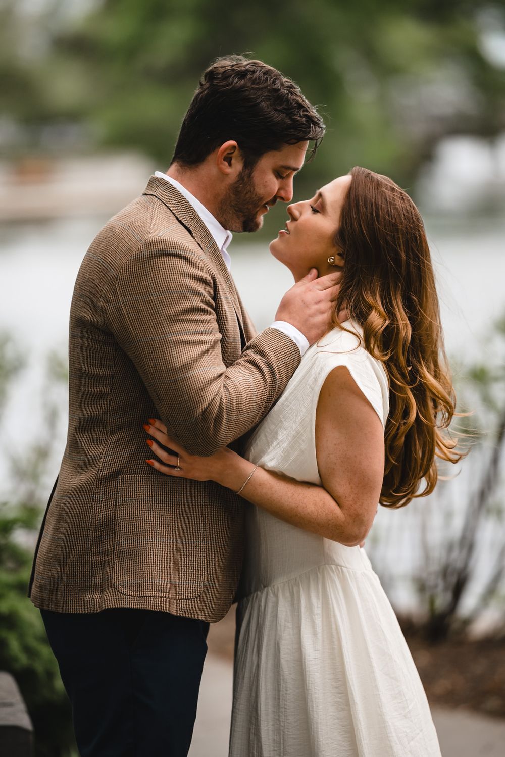 Romantic couple in elegant attire share an intimate moment outdoors, man in brown blazer gently holds woman in white dress.