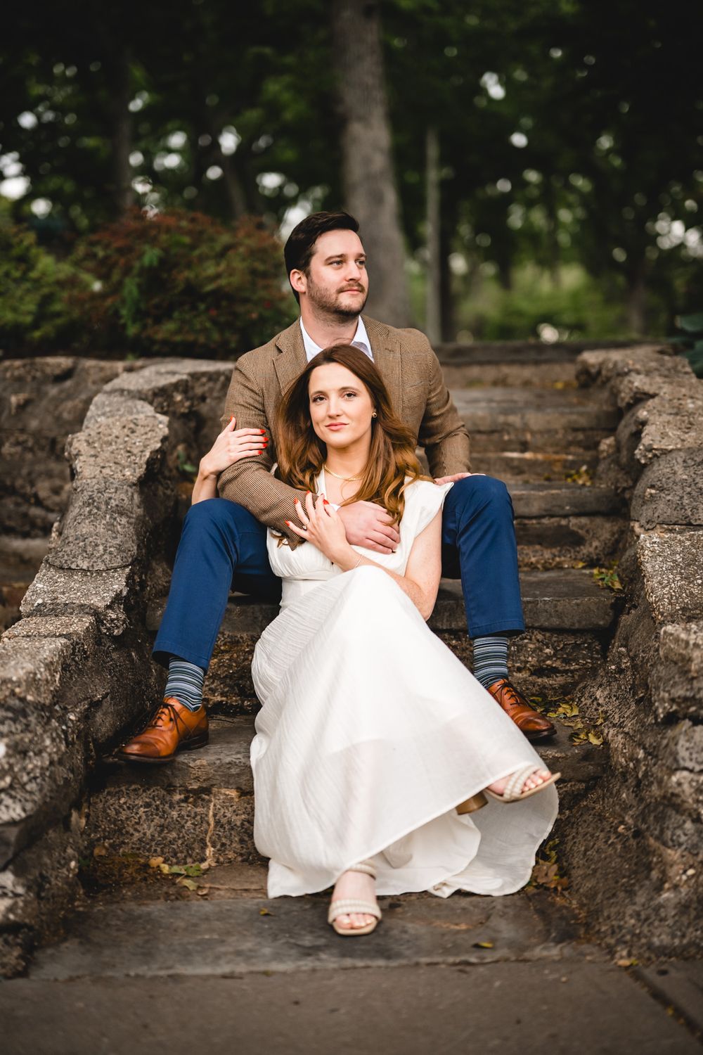 Bride and groom sitting on stone steps, woman in white dress leaning against man in brown blazer and blue pants.