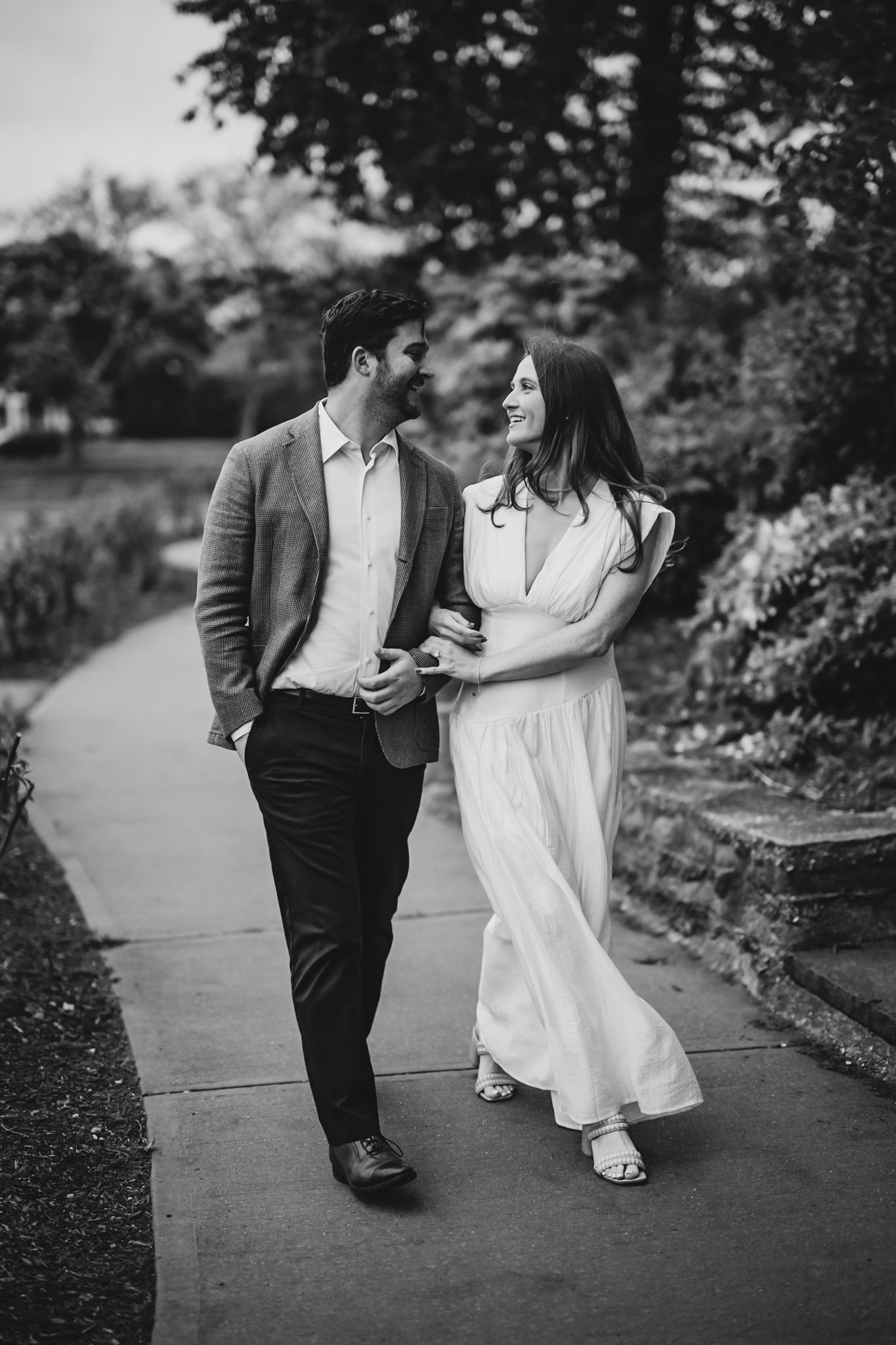 Couple walking and laughing together on a garden path, man in blazer and woman in white dress, black and white photo.