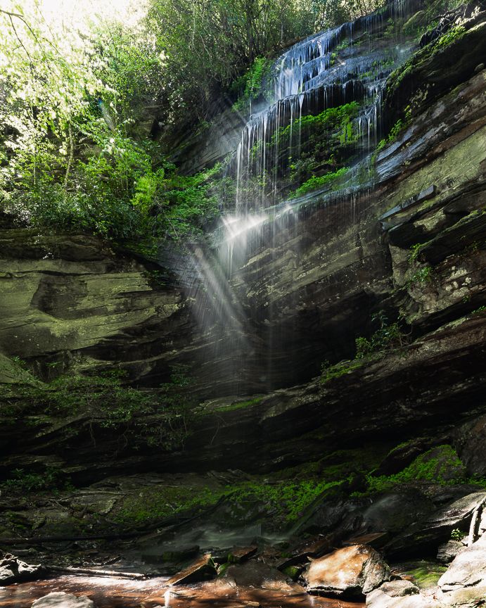 Sunlight rays illuminate a cascading waterfall over mossy rocky cliffs surrounded by lush green forest.