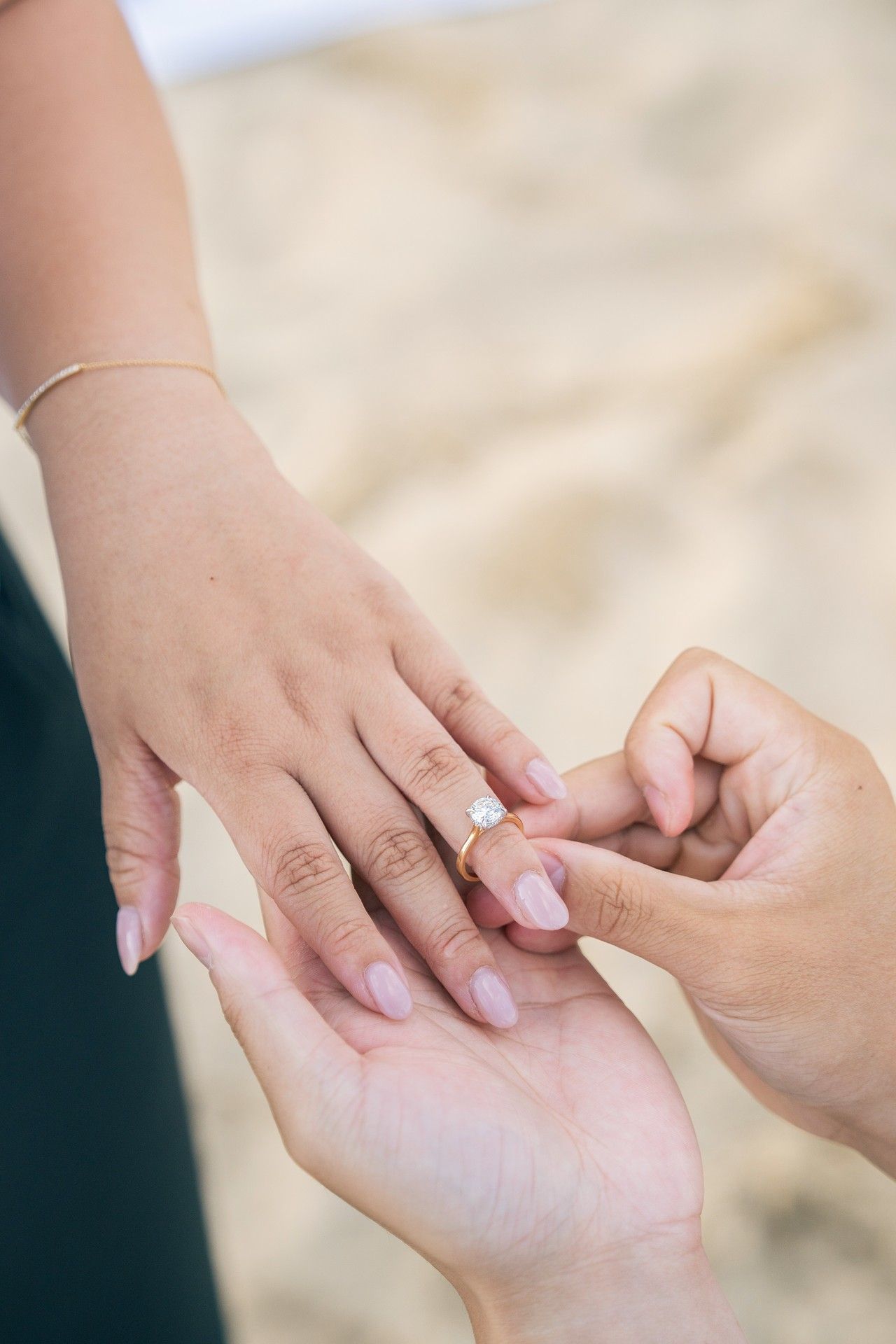 Person placing a diamond engagement ring on a woman's finger on a sandy beach during a romantic proposal.