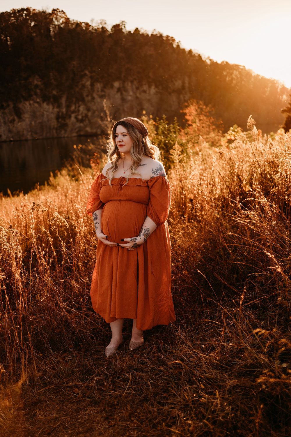 Pregnant woman in rust orange dress holding baby bump at golden hour in a field.