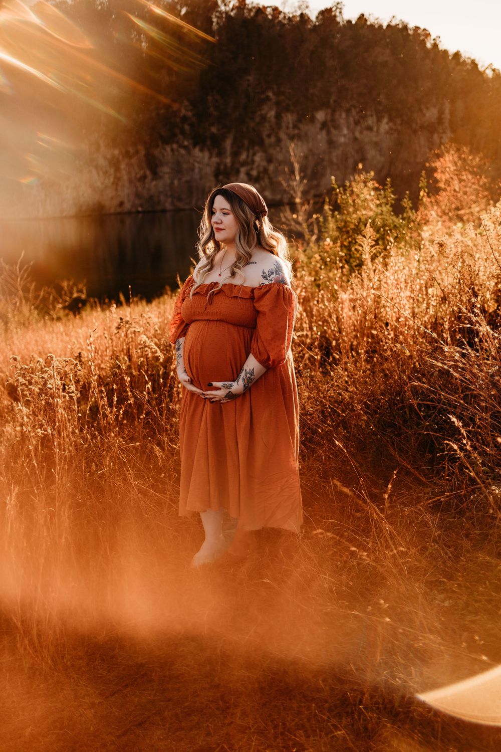 Pregnant woman in rust orange off-shoulder dress cradling baby bump in golden sunlit field.