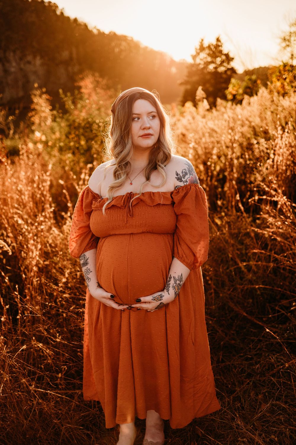 Pregnant woman in rust off-shoulder dress holding baby bump at golden hour in autumn field.