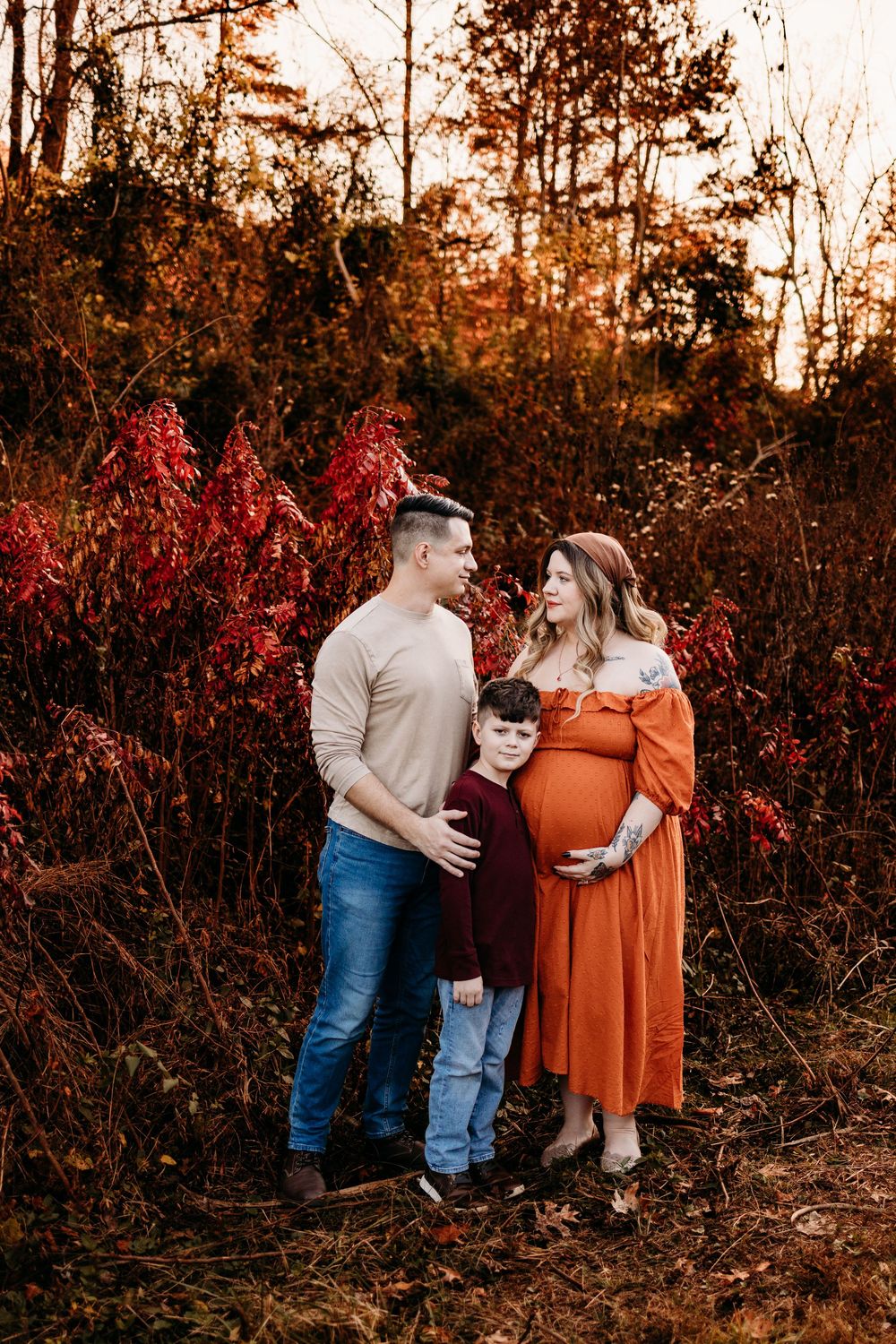 Family maternity photo with pregnant woman in orange dress, man, and child in autumn foliage setting.