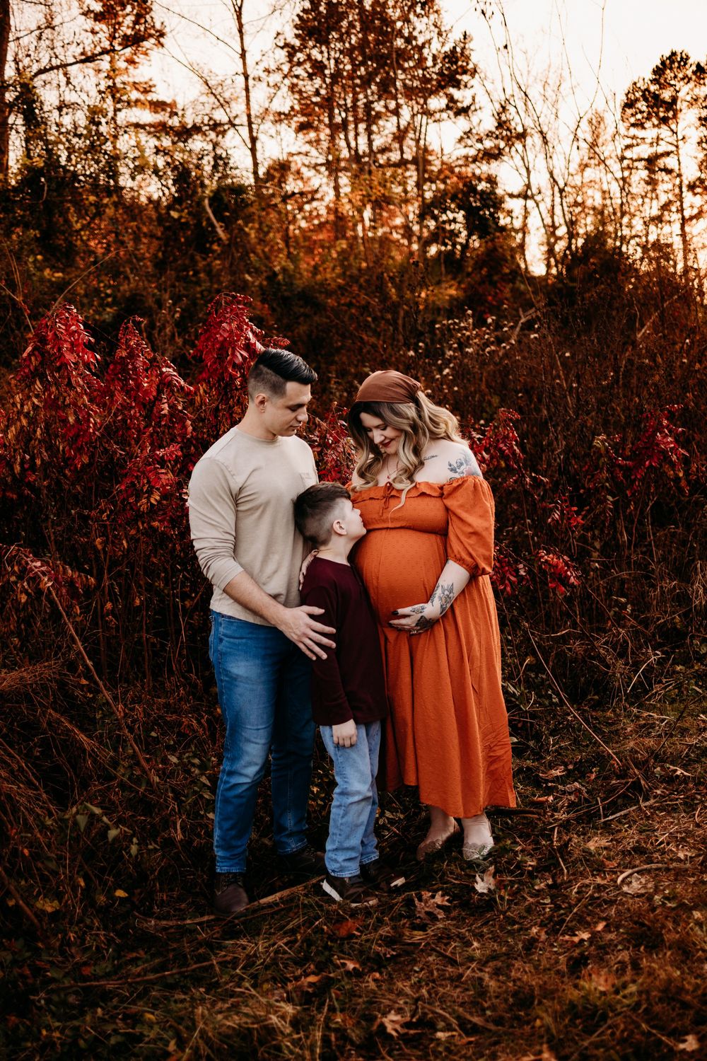 Expecting family poses outdoors in autumn, pregnant woman in orange dress with husband and young son among red fall foliage.