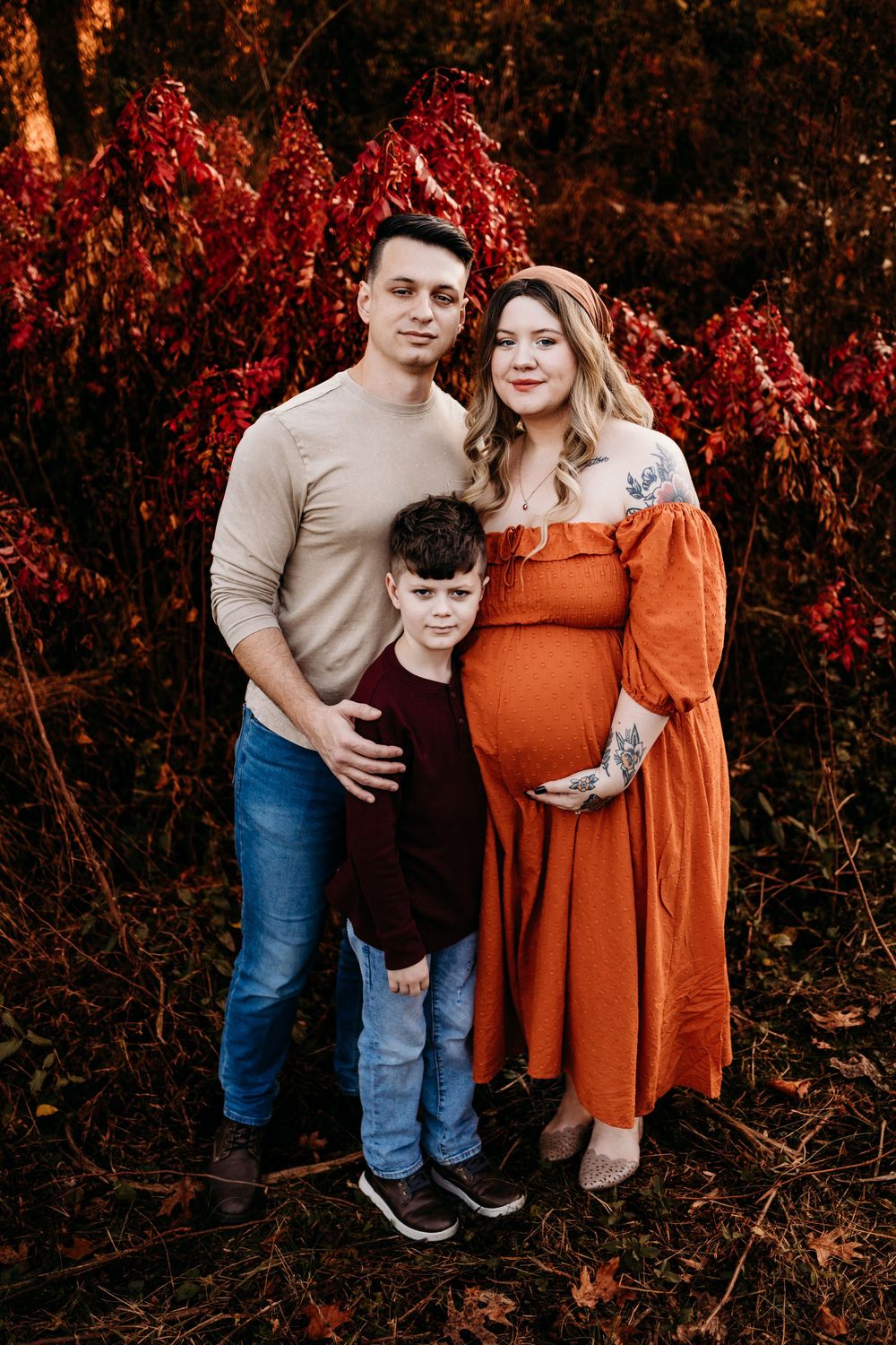 Pregnant woman in orange dress with partner and son posing in front of red autumn foliage.