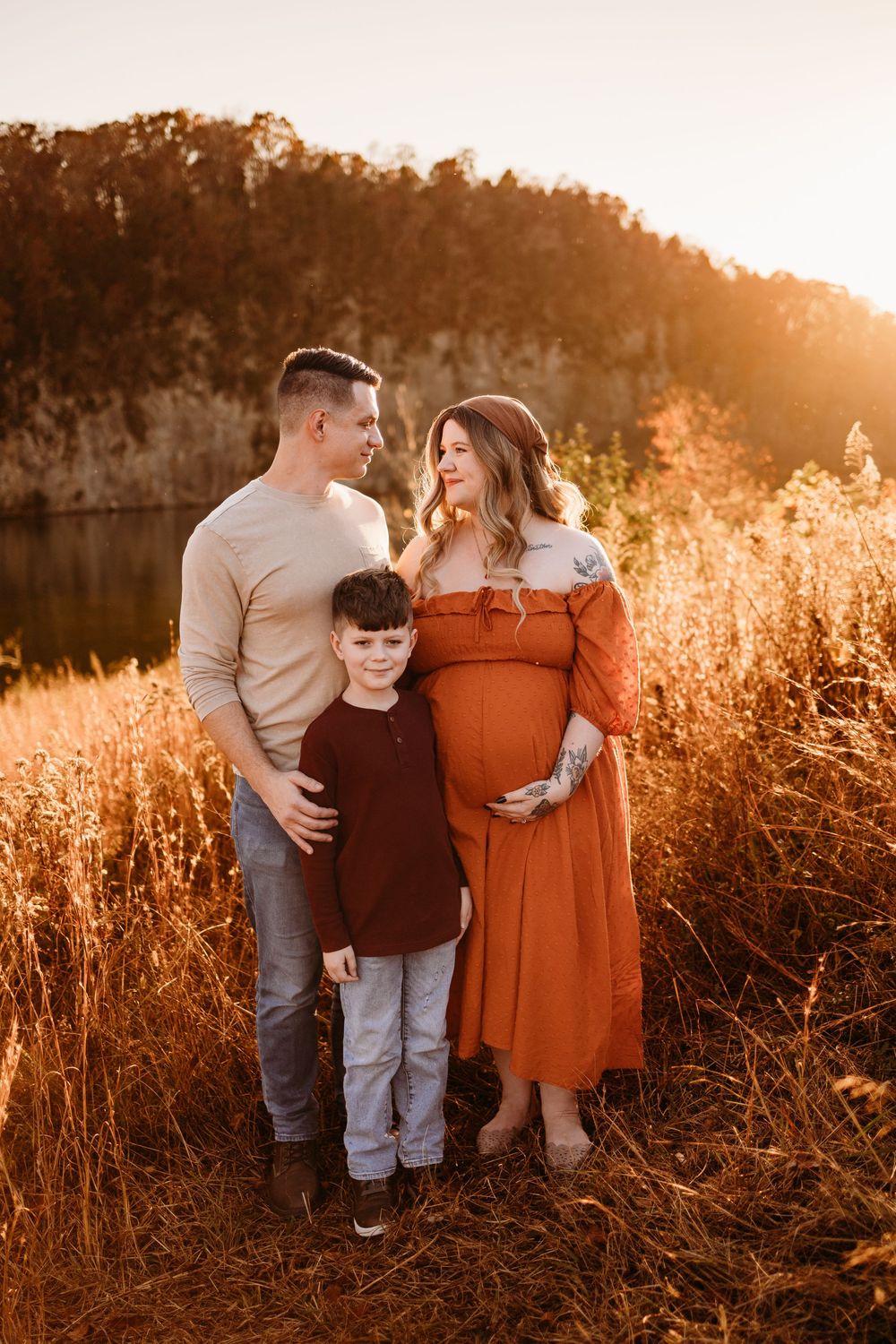 Family maternity photo at golden hour in autumn field, pregnant woman in rust dress with husband and son.