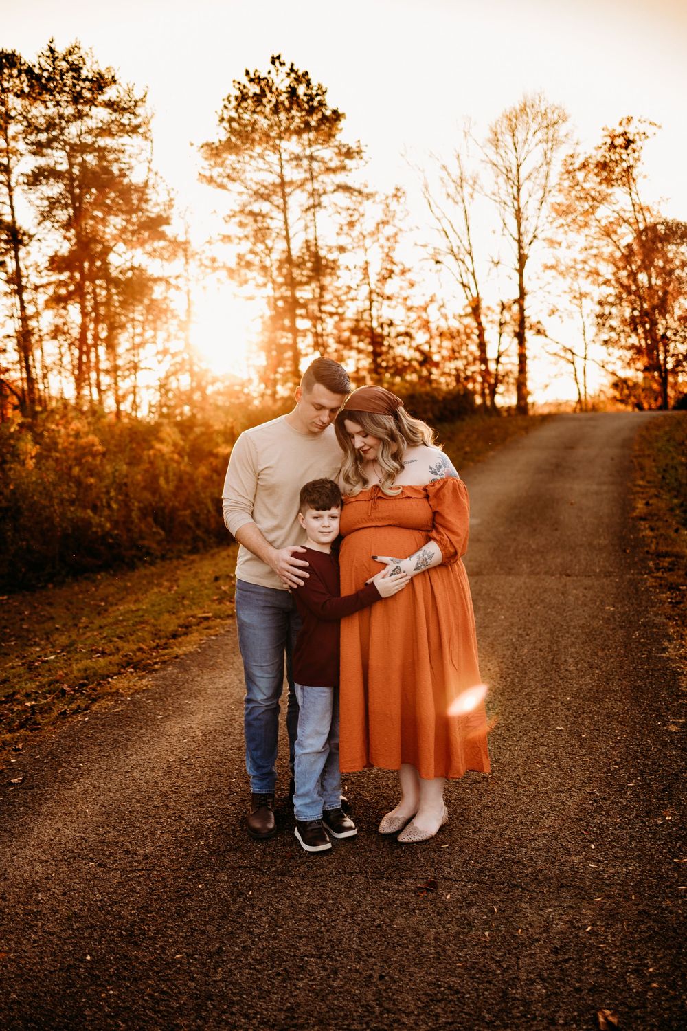 A pregnant woman in an orange dress poses with her husband and son at golden hour on a dirt road.