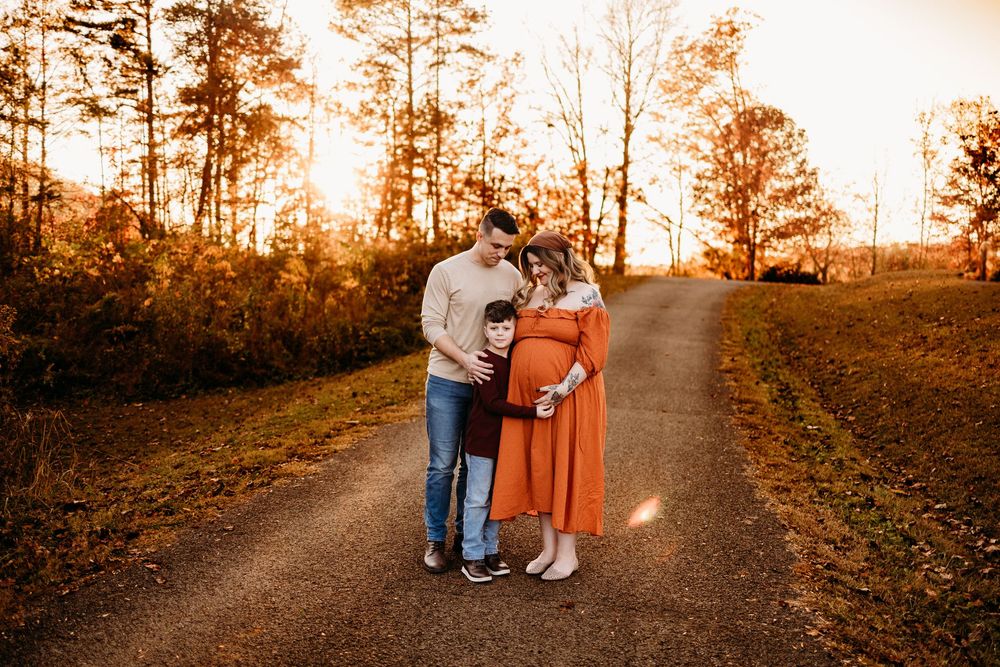A family of three embraces on a scenic dirt road surrounded by fall foliage at sunset.