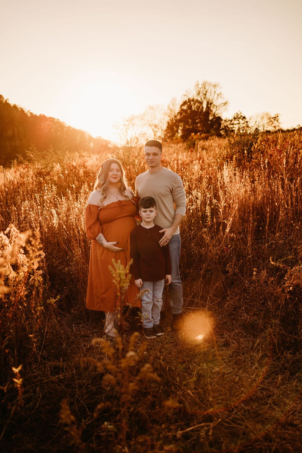 Pregnant woman in rust dress poses with husband and son in golden field during sunset maternity photo session.